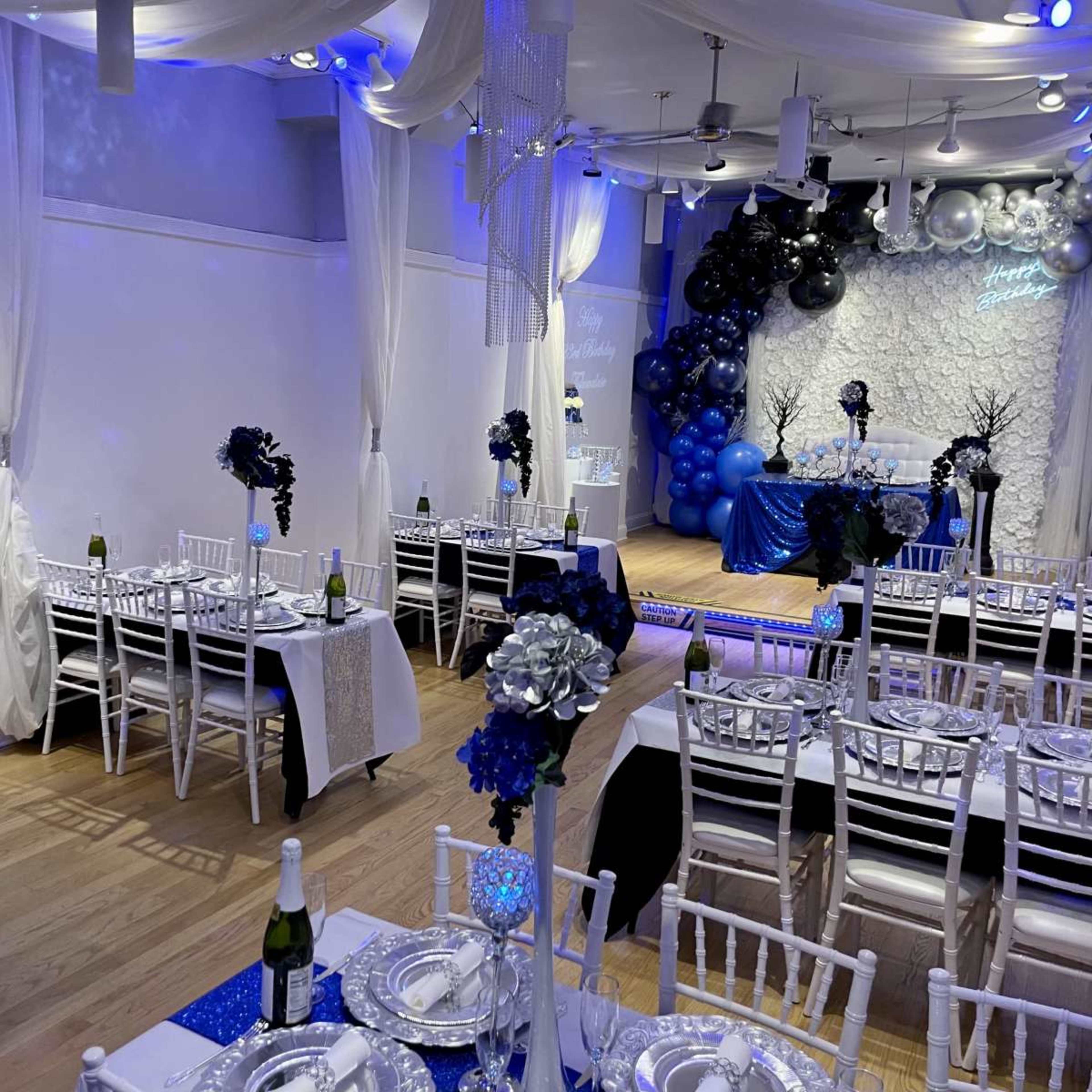 The image shows a decorated event space with tables set for a celebration, featuring blue and silver balloons, floral centerpieces, and white chairs.