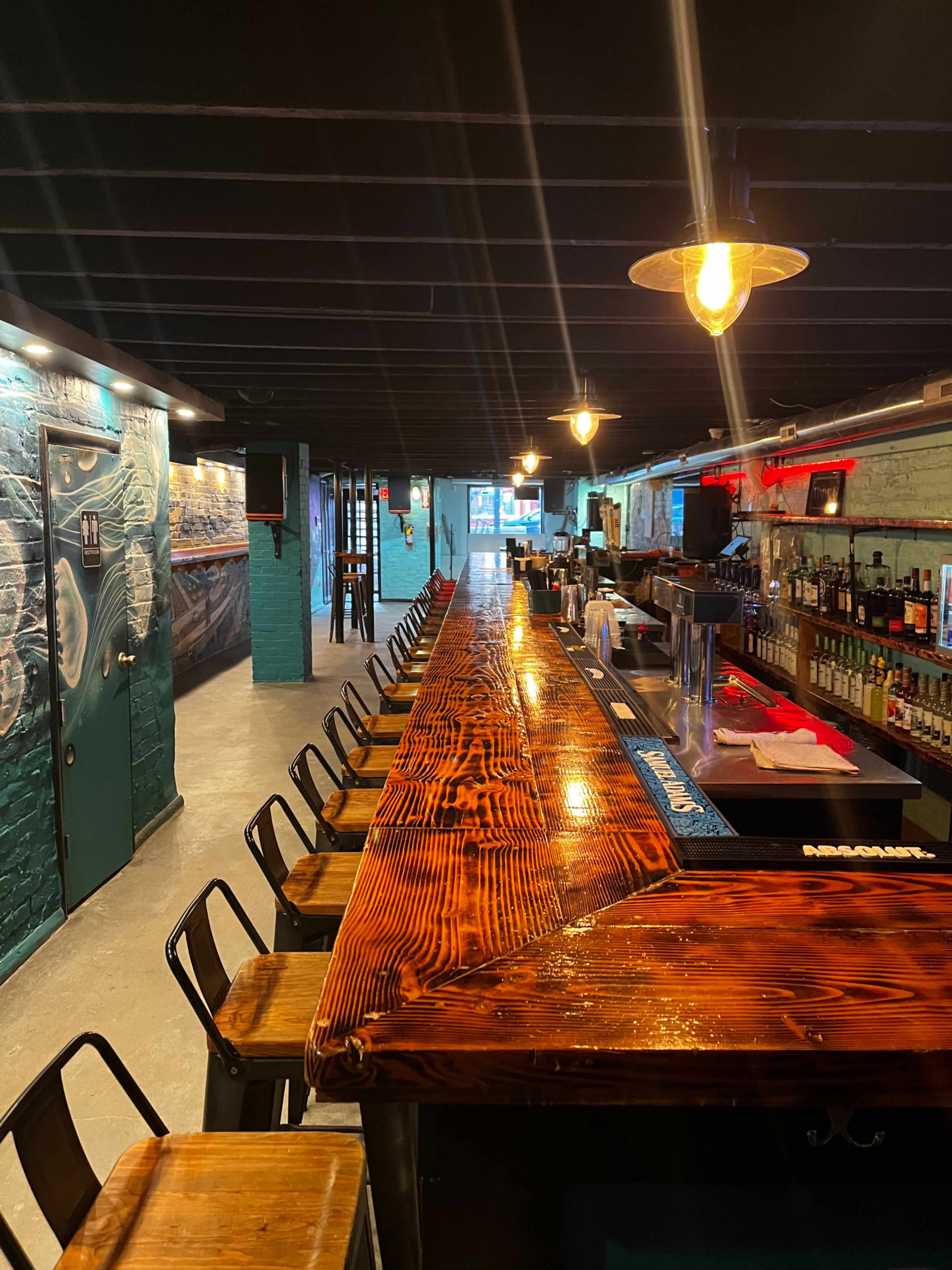 A dimly lit bar with a long wooden countertop and several stools lined up along one side, leading to a well-stocked bar area in the background.