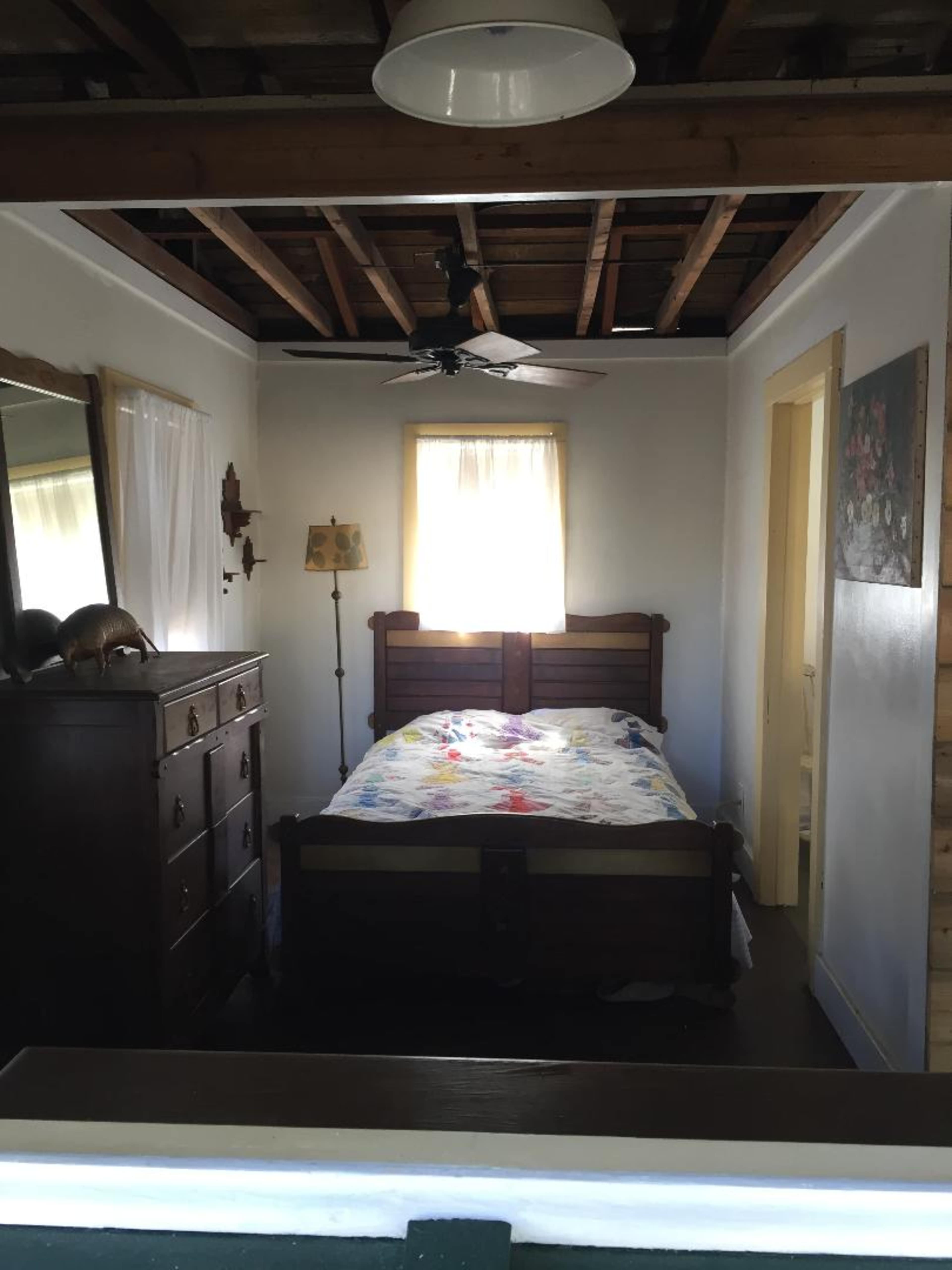 The image shows a simple bedroom with a wooden bed, a dresser, a lamp, and a window with natural light filtering in.
