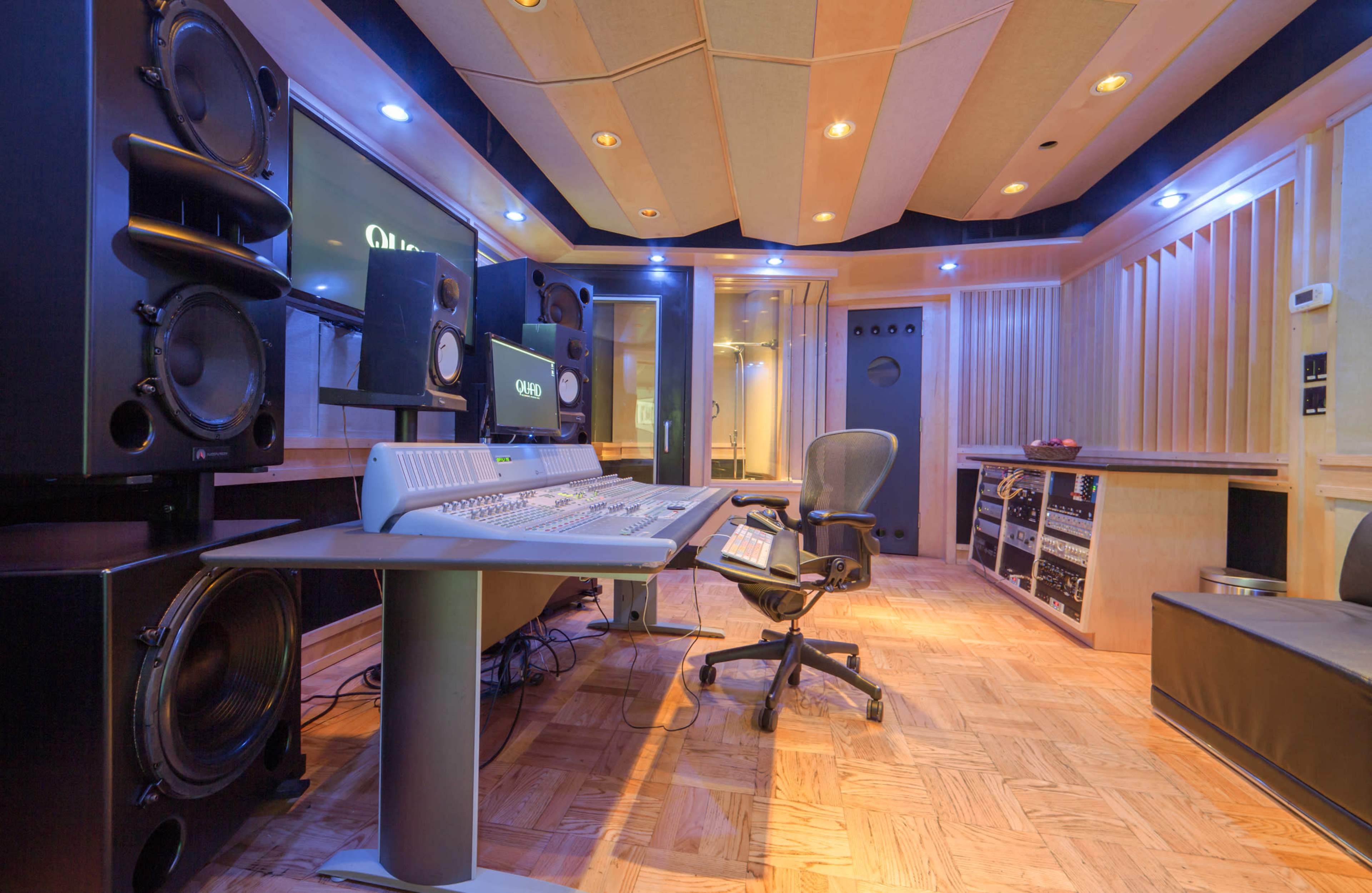 The image shows a modern audio recording studio featuring a large sound mixing console, high-quality speakers, and acoustic paneling on the walls.