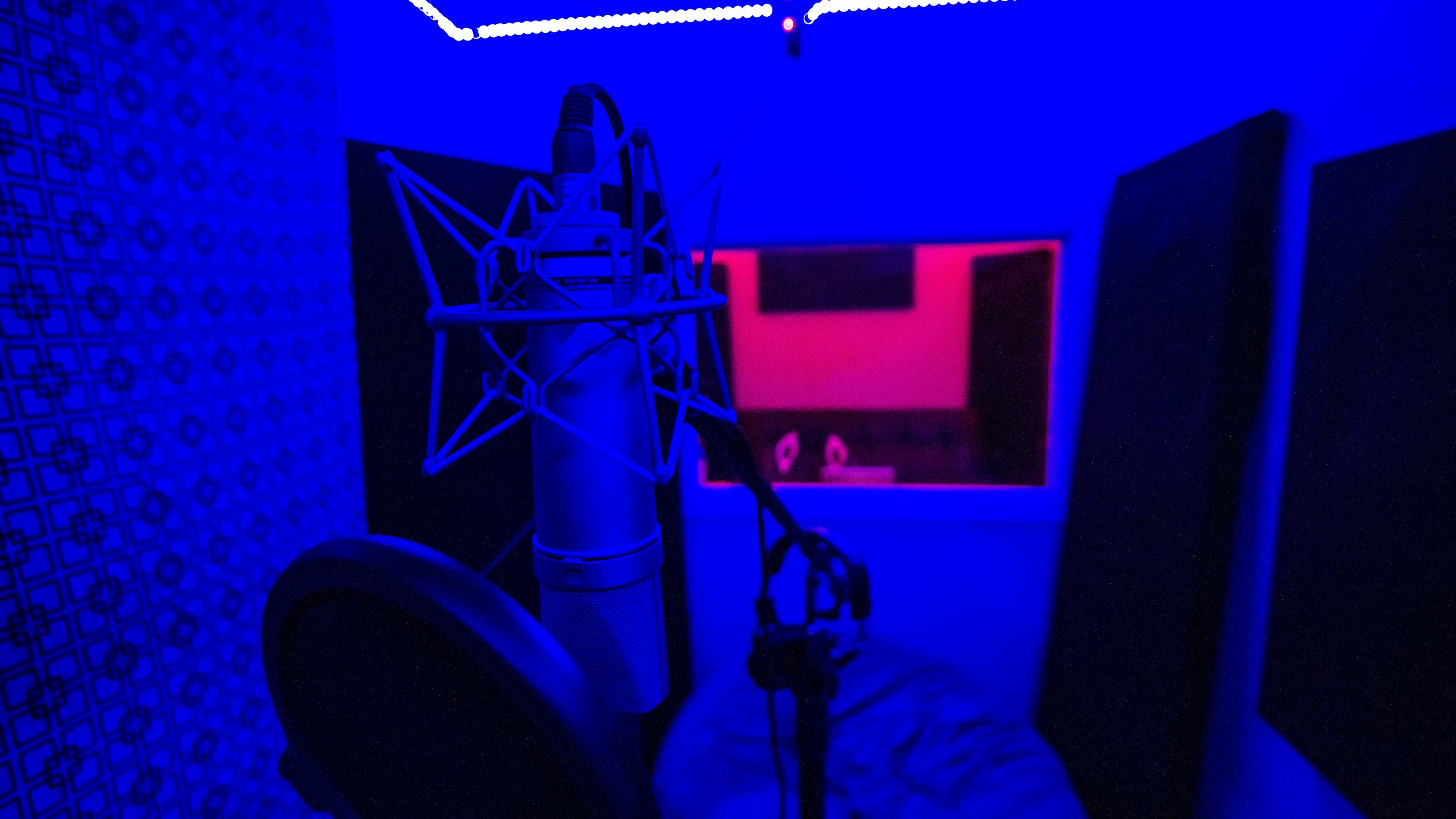 A recording studio with a microphone in the foreground and a control room visible through a window, all illuminated by blue and red lights.
