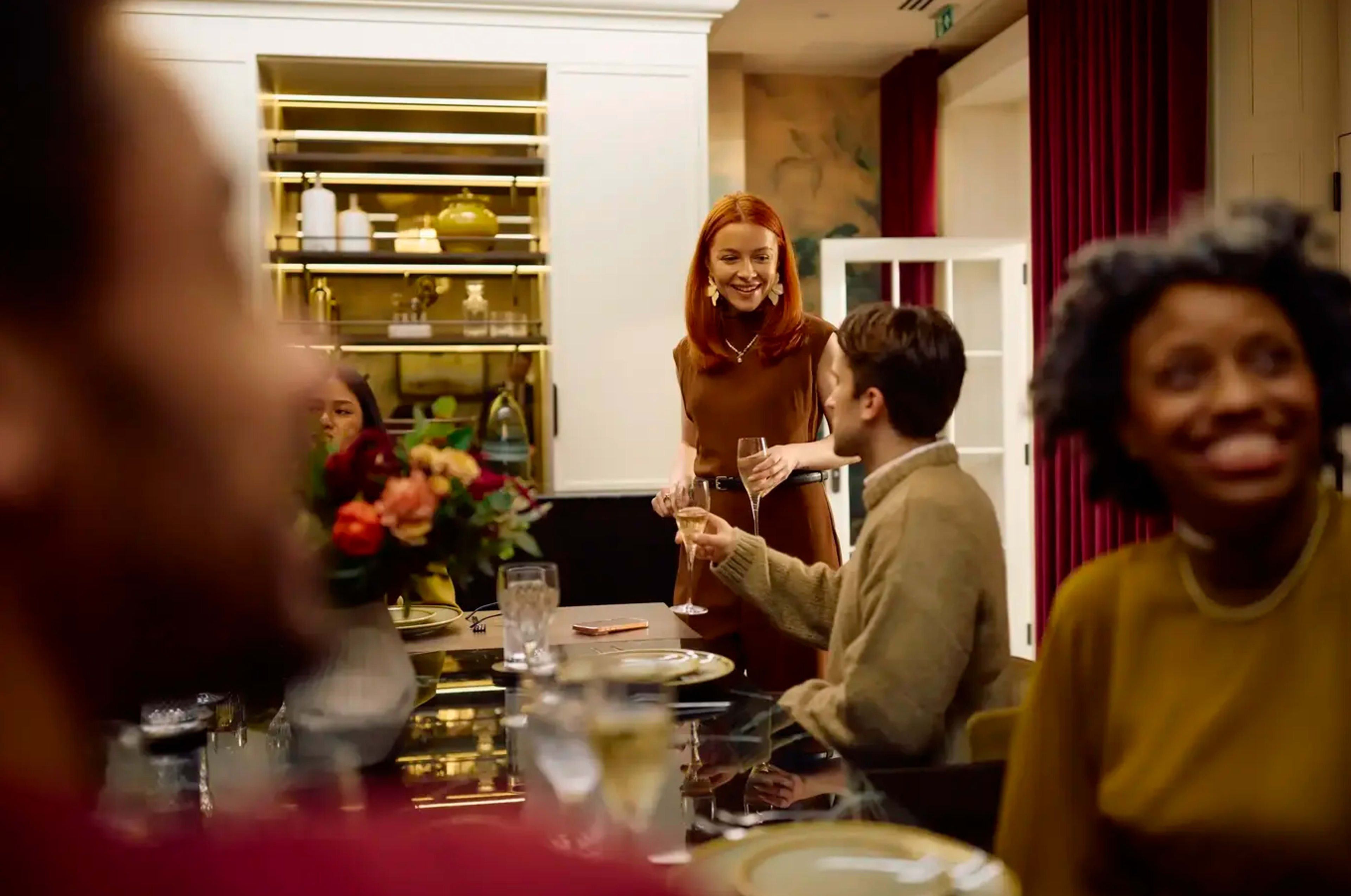 A group of people is gathered around a dining table, smiling and engaging in conversation while one woman stands and holds a drink.