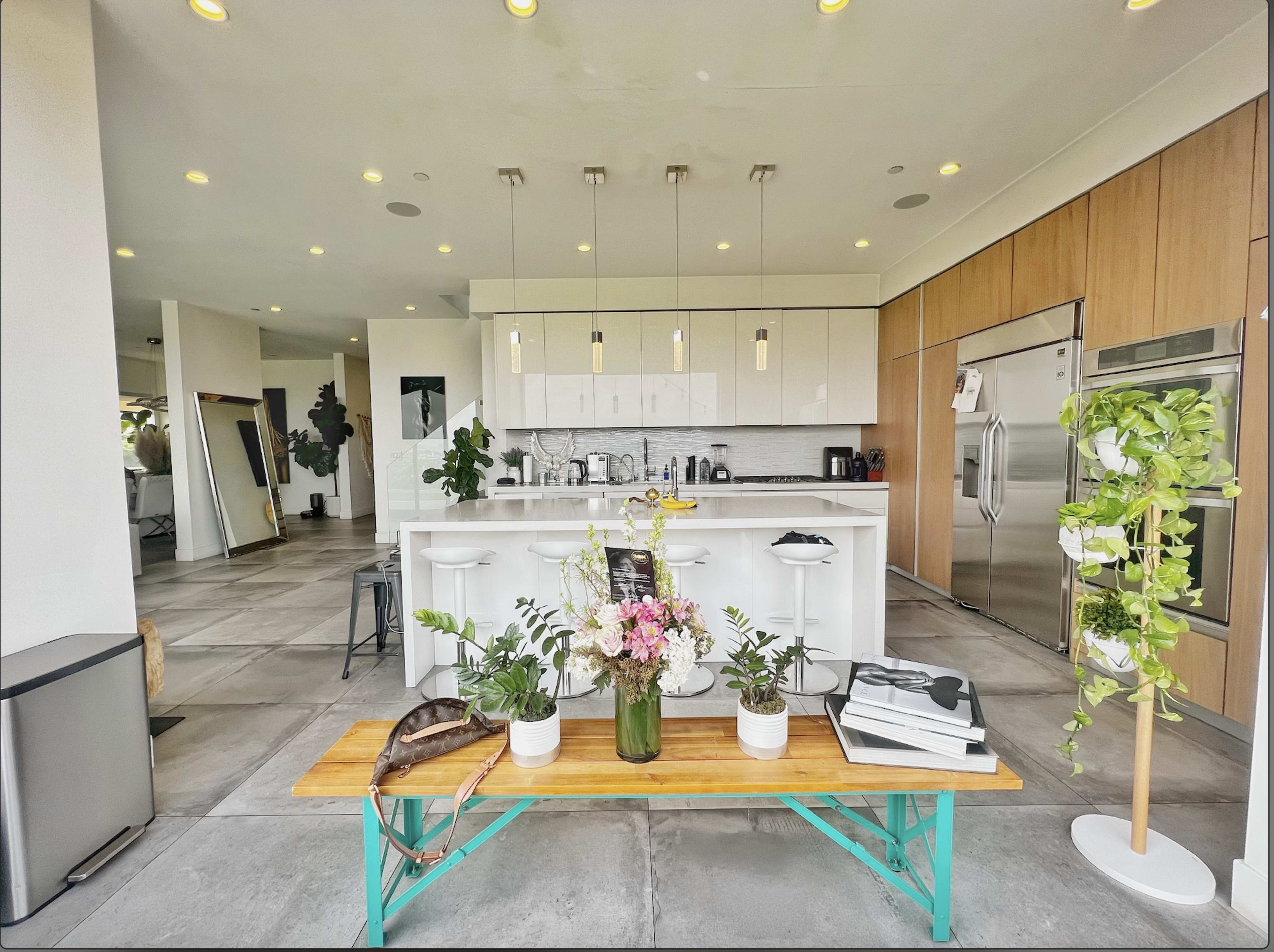 A modern kitchen and dining area with a wooden table in the foreground adorned with flowers and a few decorative items, while sleek cabinetry and stainless steel appliances are visible in the background.