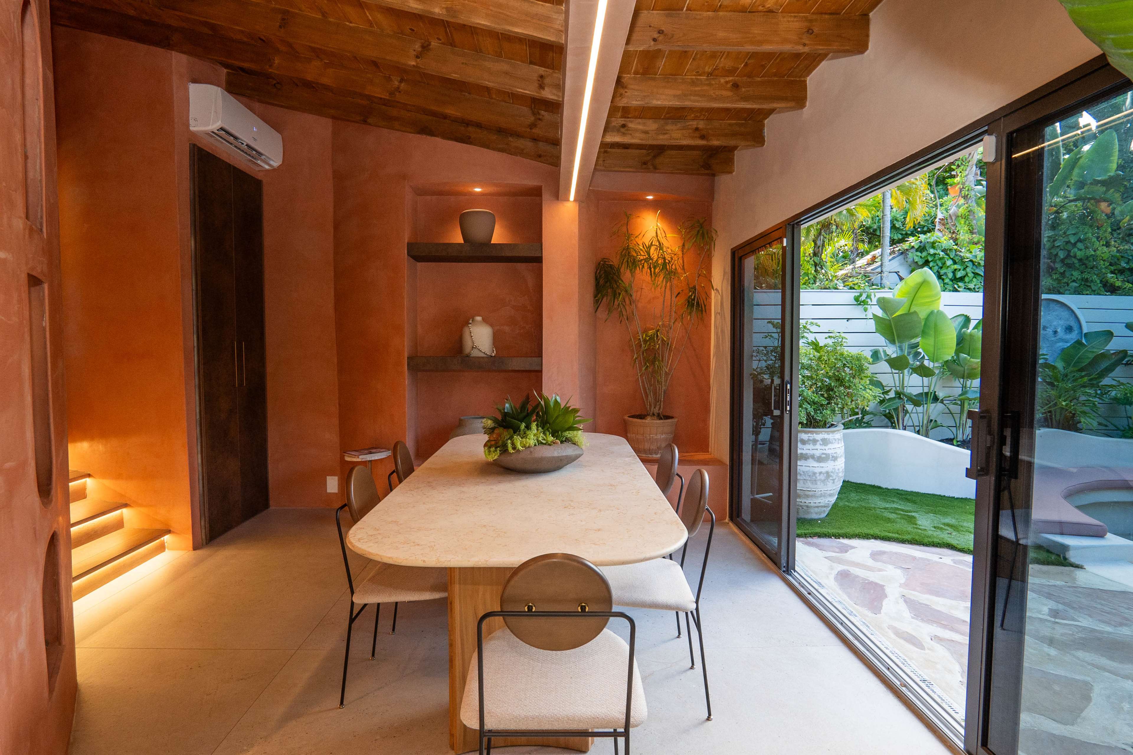 The image shows a modern dining area featuring a stone table surrounded by minimalist chairs, with an open view of a lush outdoor space through large glass doors.