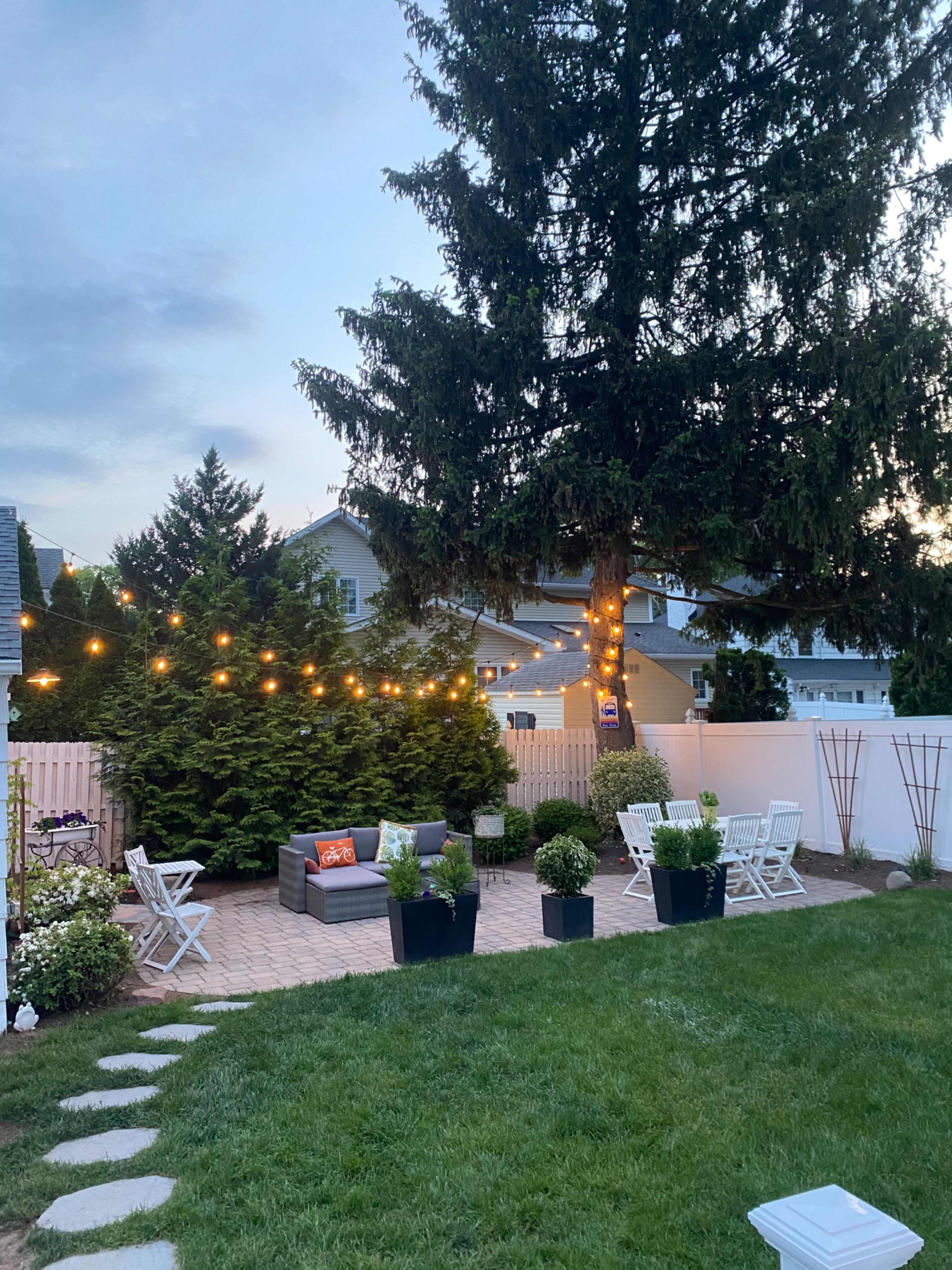 A backyard patio area with string lights overhead, a seating arrangement on a stone patio, and potted plants along the perimeter.