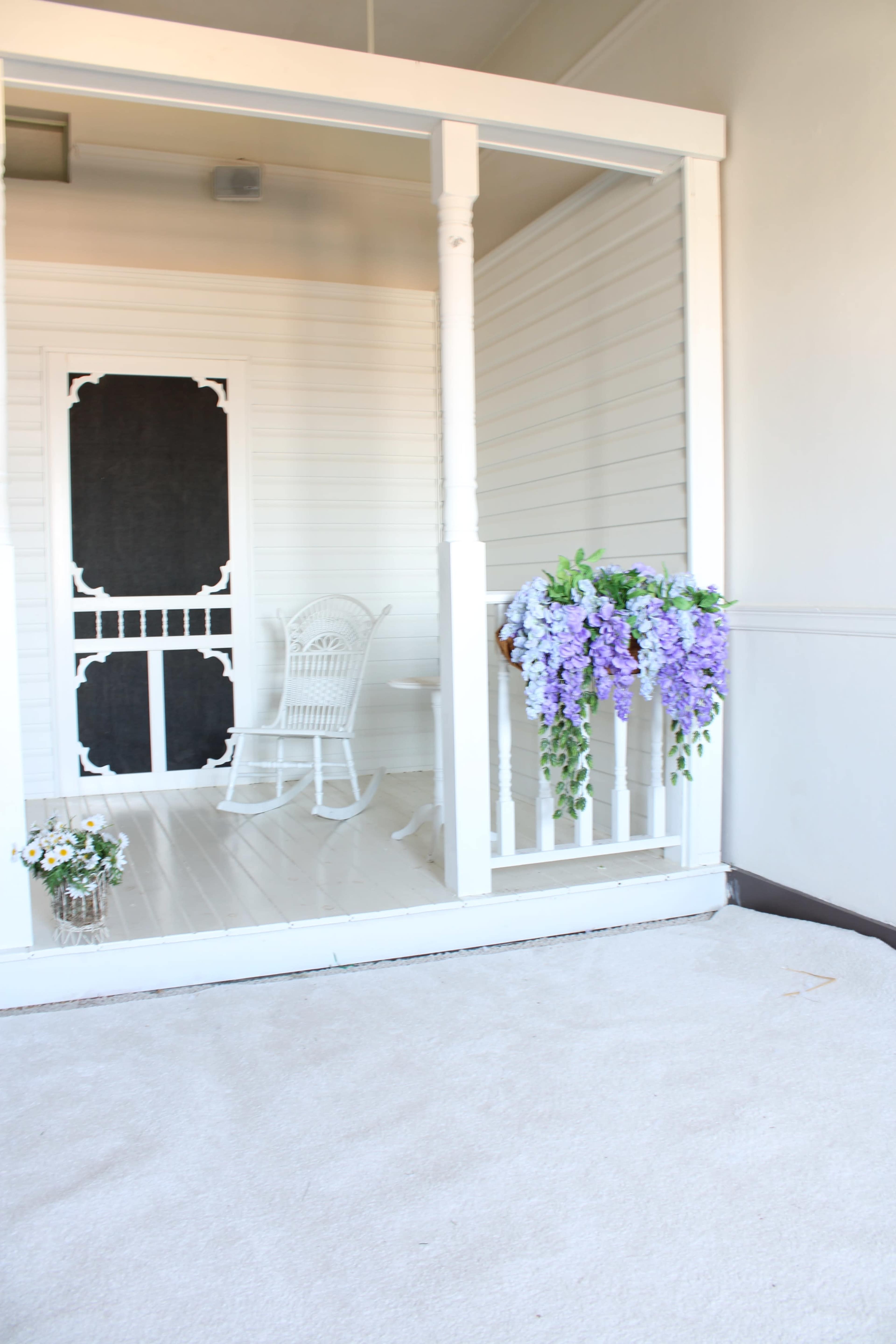 The image shows a porch with a white rocking chair, a decorative black door, and hanging flowers in shades of purple.