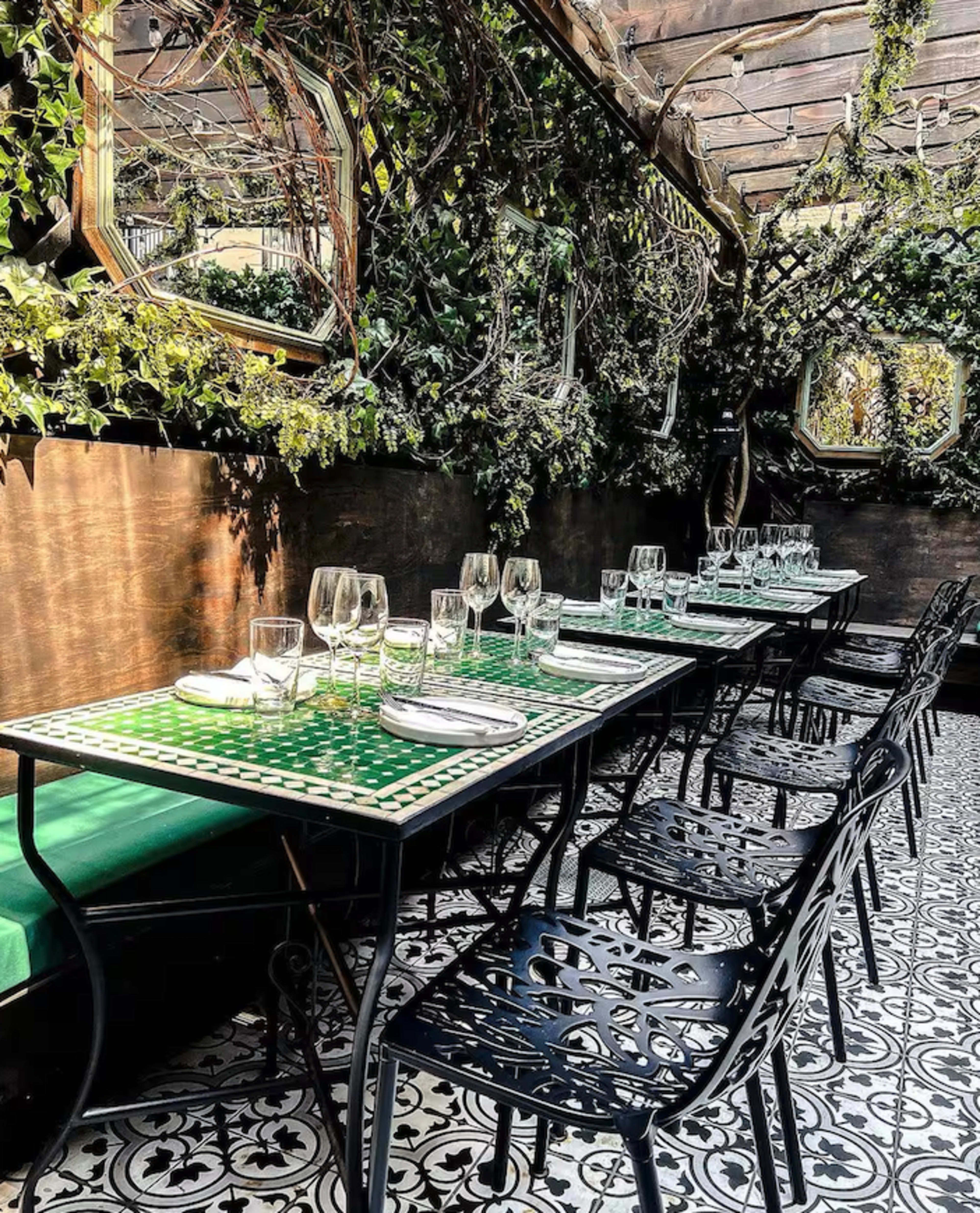 A long table set for dining with glassware and white napkins is placed under a canopy of greenery in a decorated restaurant space.