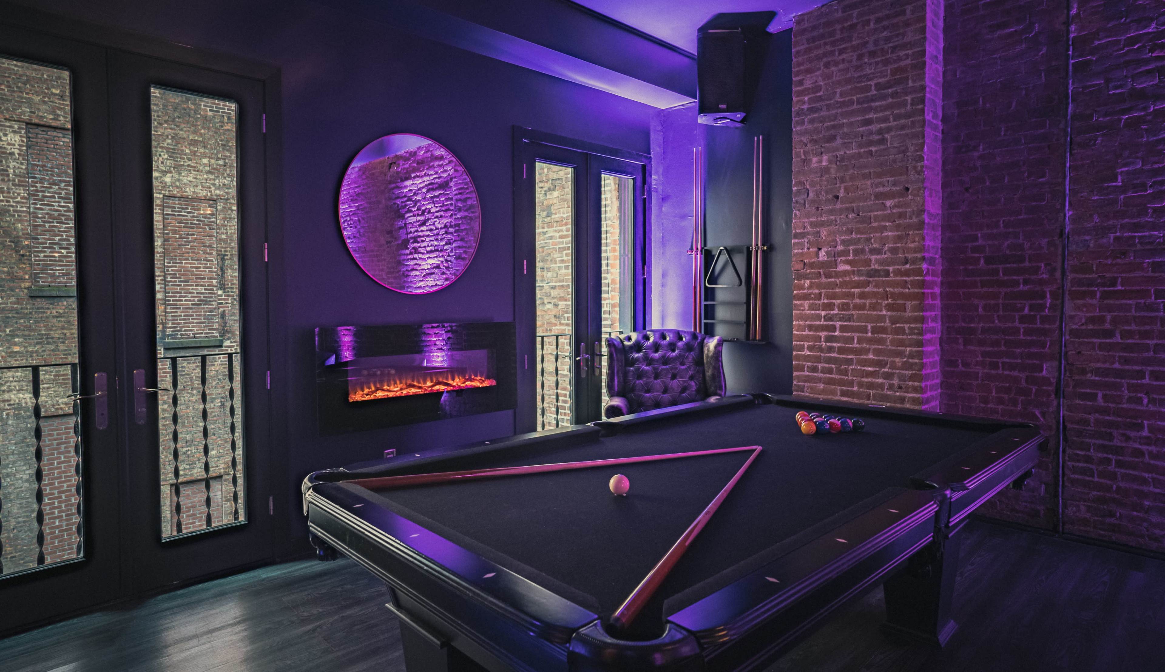 The image shows a game room with a pool table, brick walls, and purple lighting, featuring a modern fireplace and large windows.