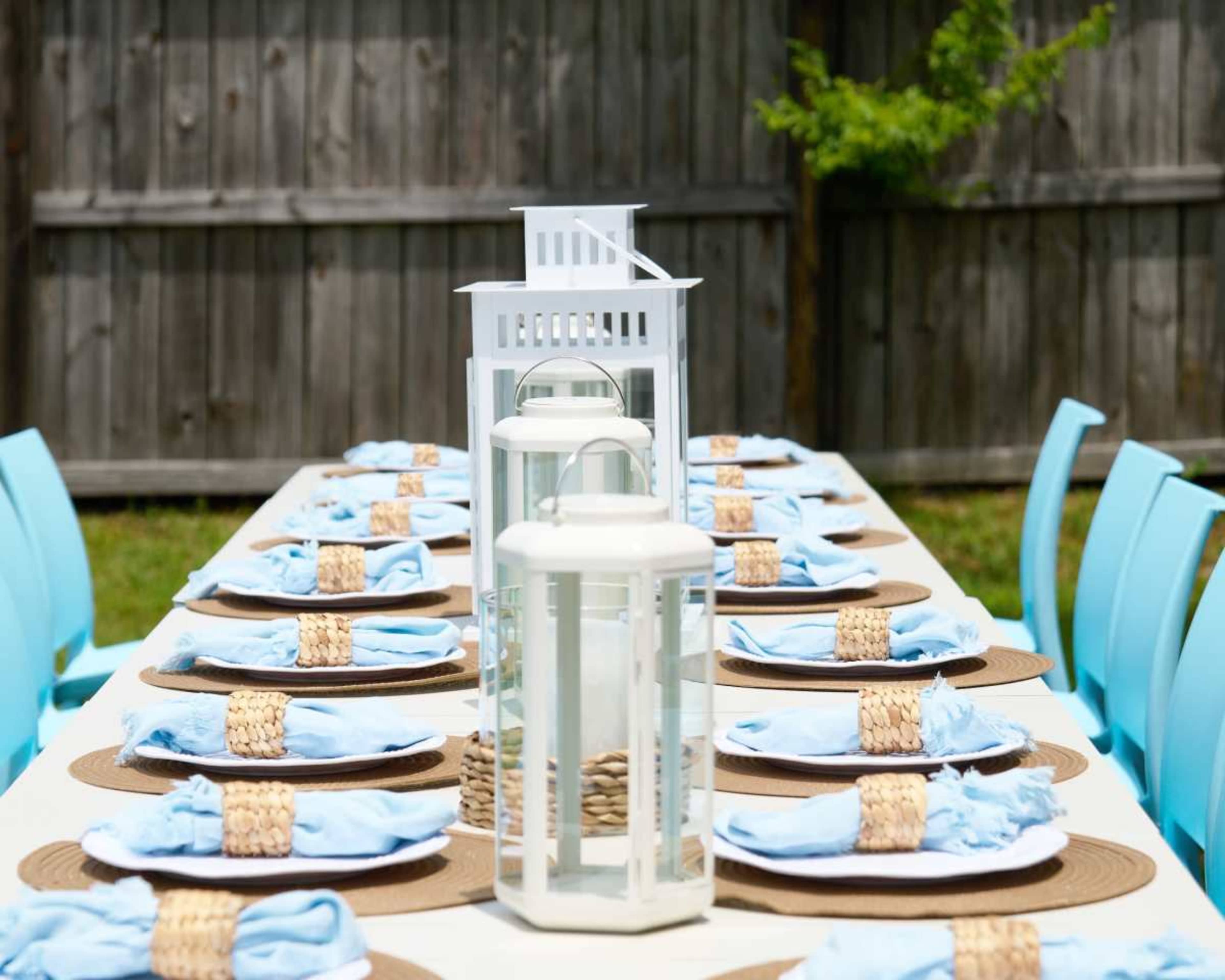 A long outdoor dining table is set with blue napkins, wicker chargers, and white lanterns, surrounded by aqua chairs against a wooden fence.