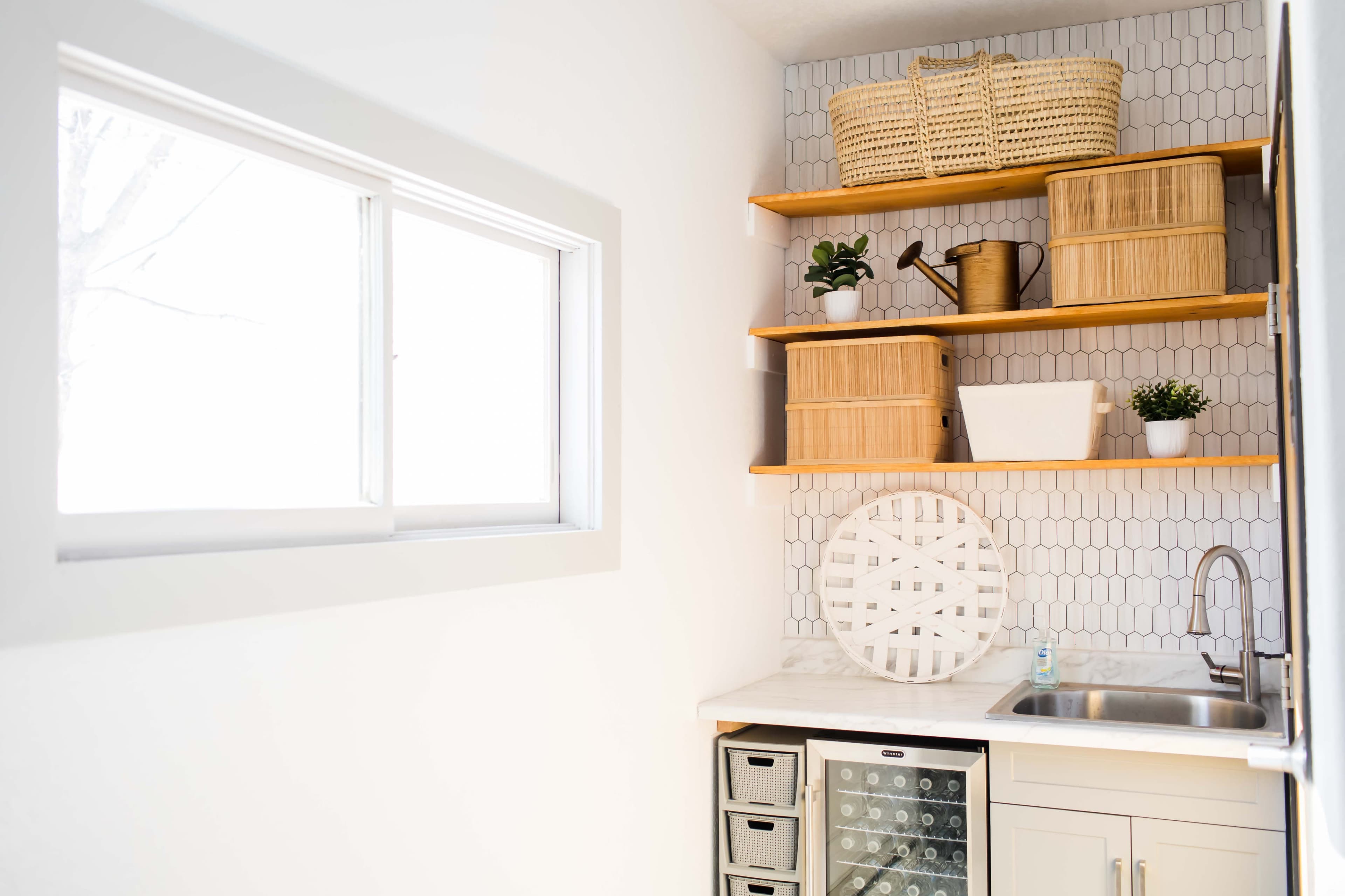 A minimalist kitchen corner featuring a window, open shelves with woven baskets, a sink, and a small refrigerator beneath the counter.