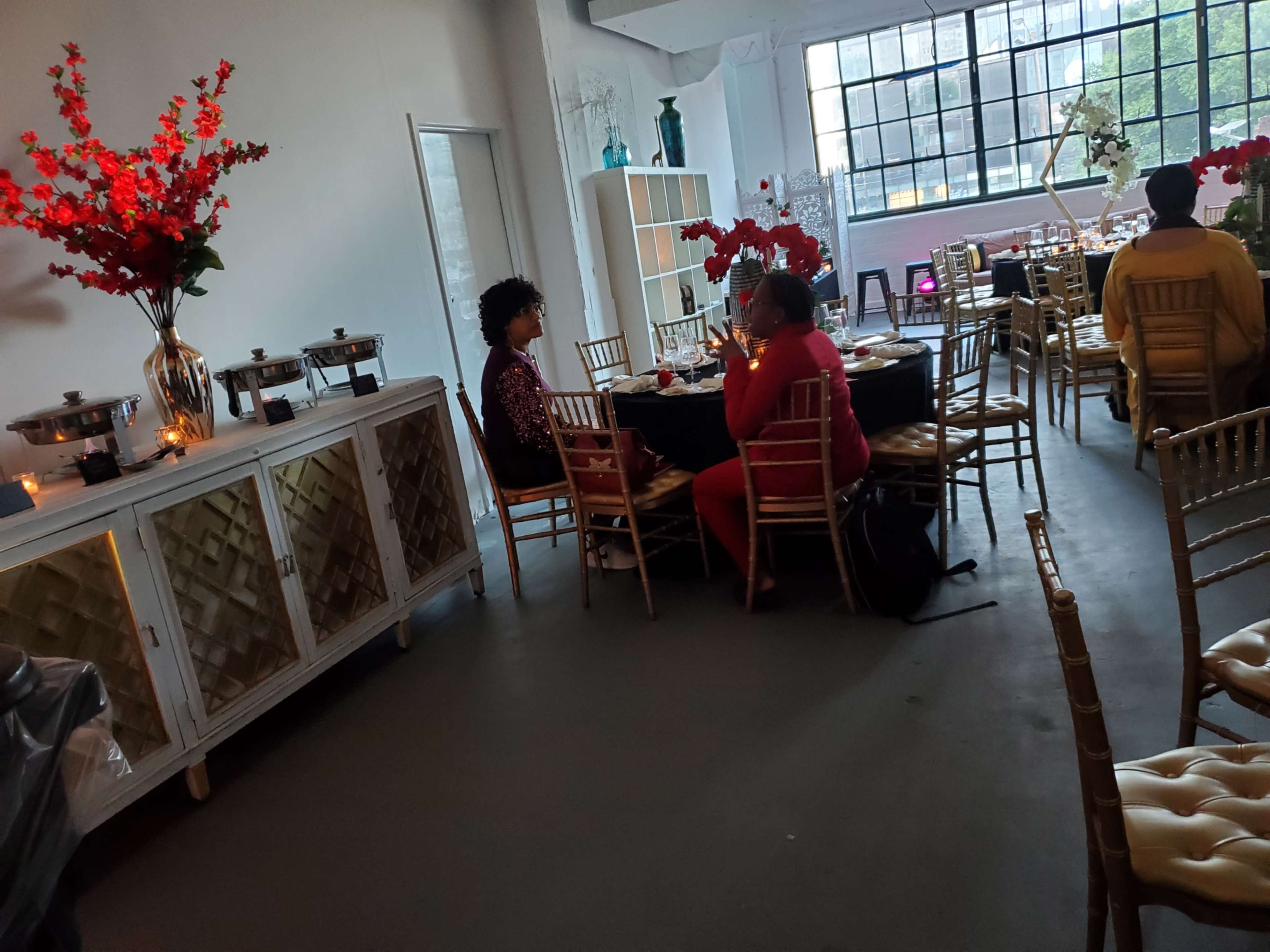 Two people are seated at a table in a decorated dining area with floral arrangements and buffet trays.