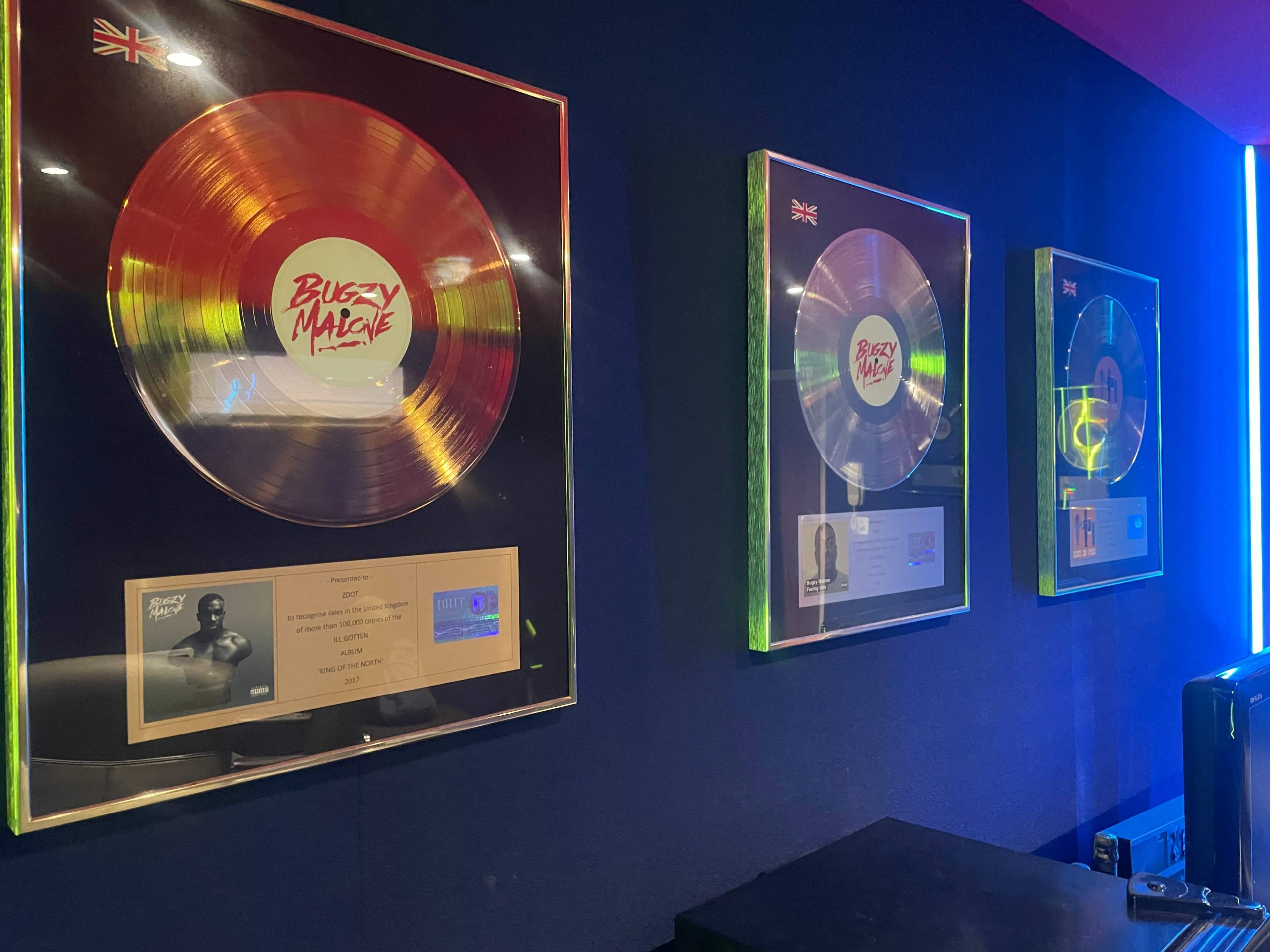 The image shows three framed music awards displayed on a blue wall, with the leftmost award featuring a gold record and a title that includes "Bugzy Malone."