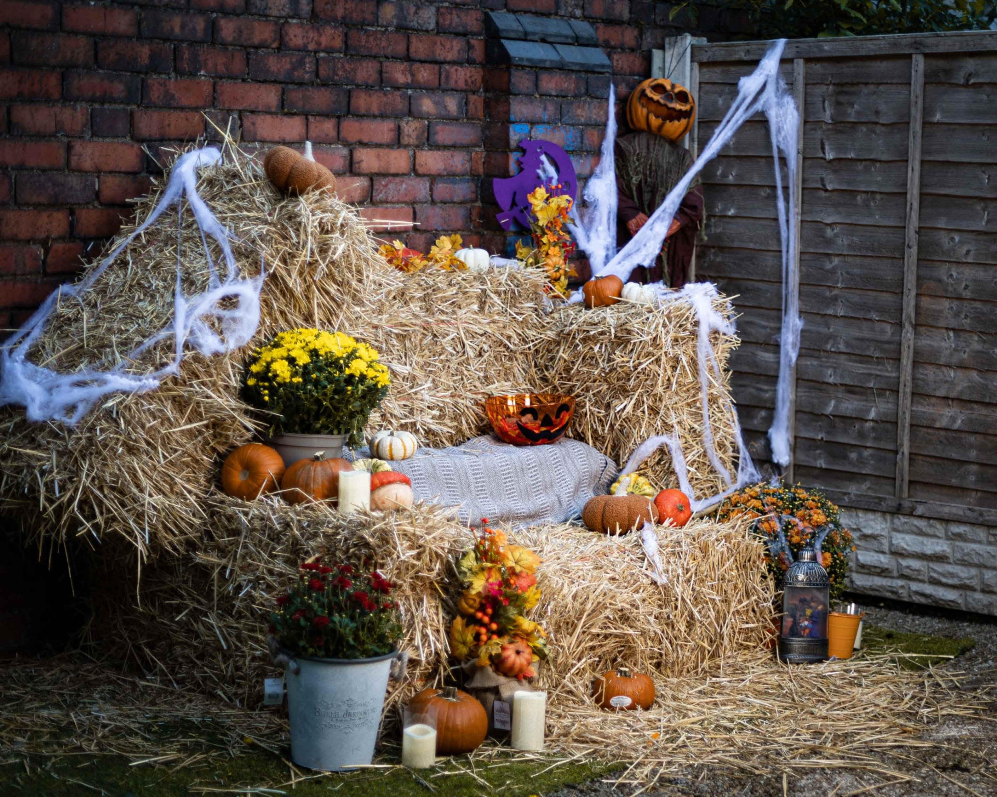 The image depicts a cozy outdoor seating area made of hay bales, decorated with pumpkins, flowers, candles, and Halloween-themed accents.