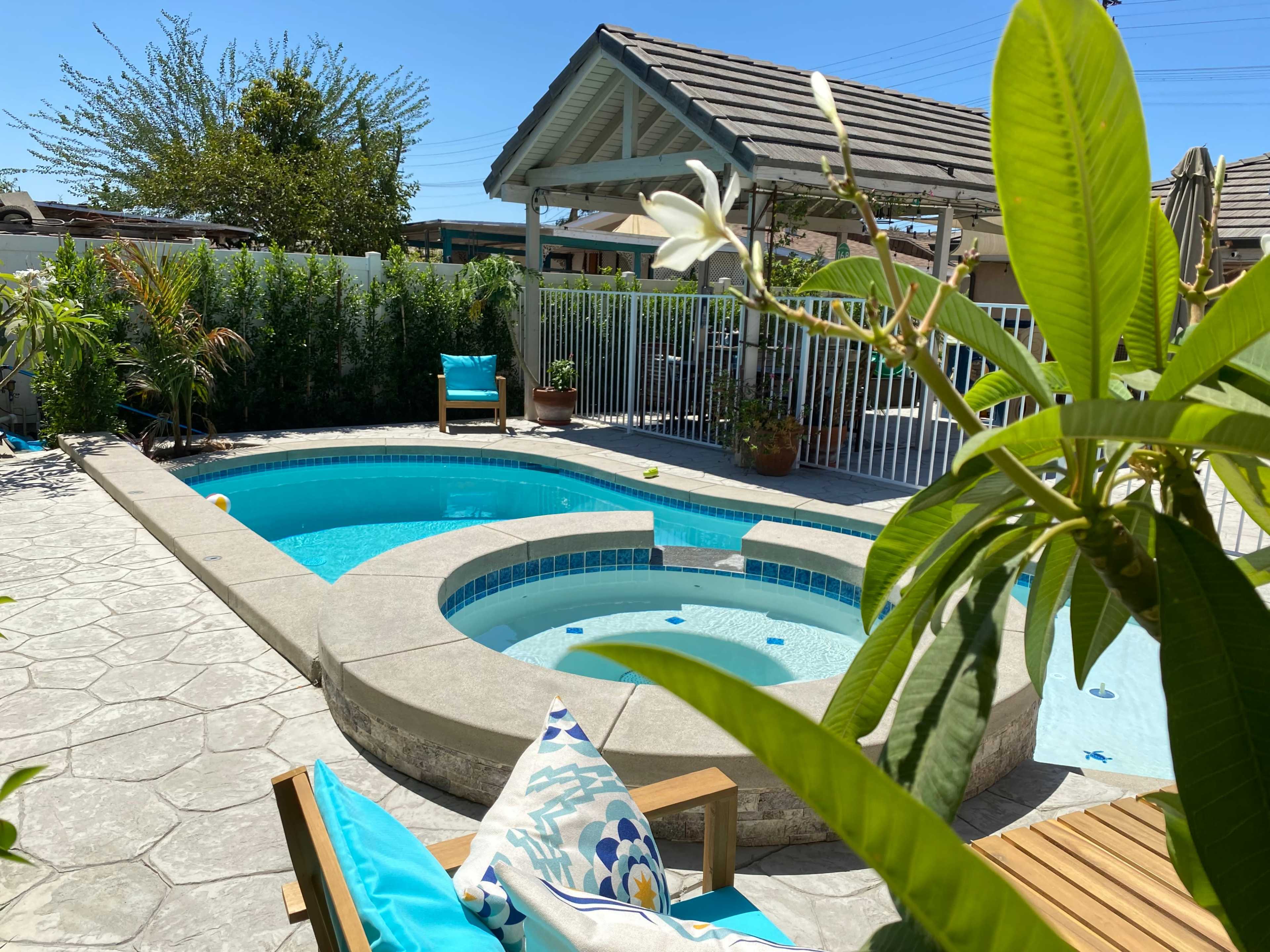 A backyard area features a pool and spa surrounded by plants, with a wooden lounge chair and a covered seating area in the background.