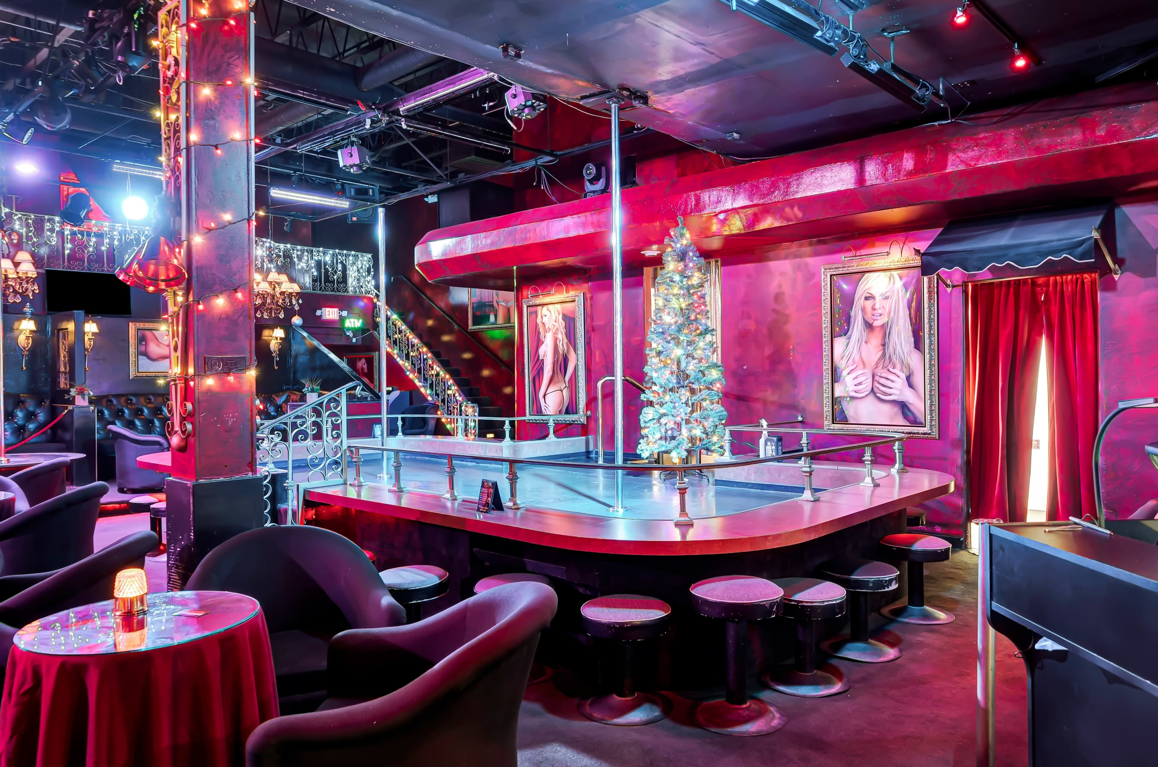 The image shows a dimly lit bar or club interior with round tables, black stools, and a stage framed by decorative lighting and artwork on the walls.