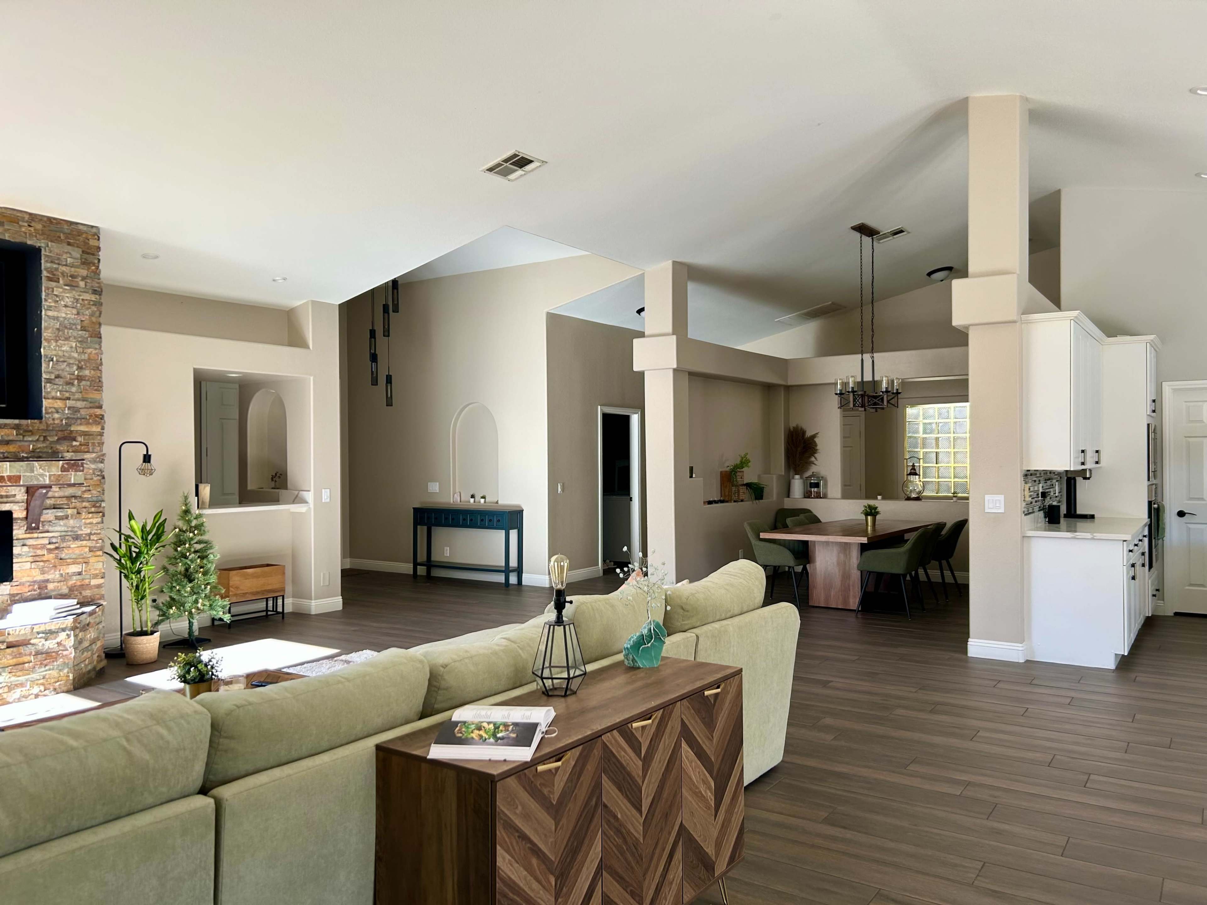A spacious living room features a green sectional sofa, a wooden side table, and an open layout leading to a dining area and kitchen.