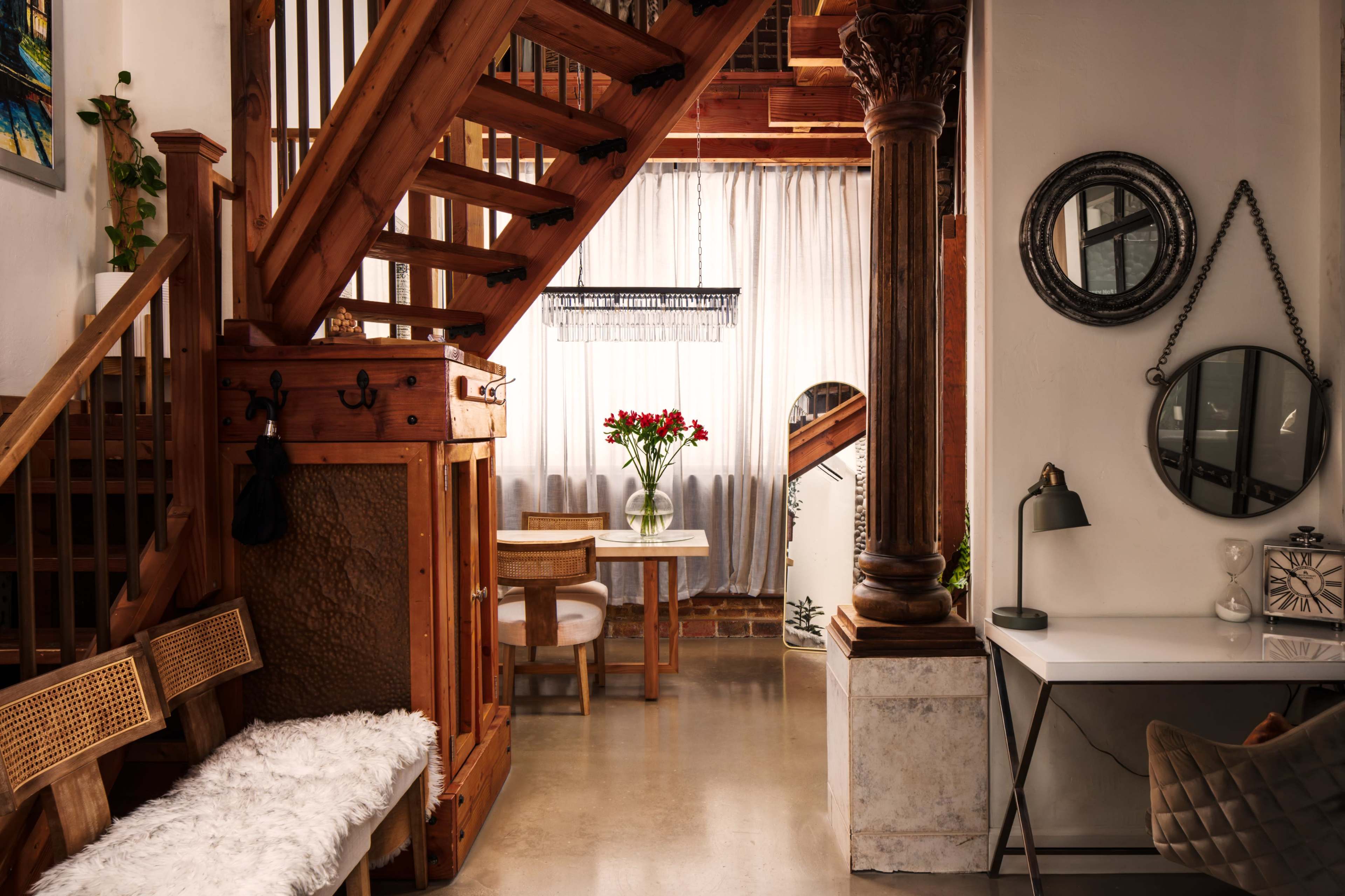The image shows a well-lit interior space featuring a wooden staircase, a small dining table with flowers, and various decorative elements including a mirror and a lamp.