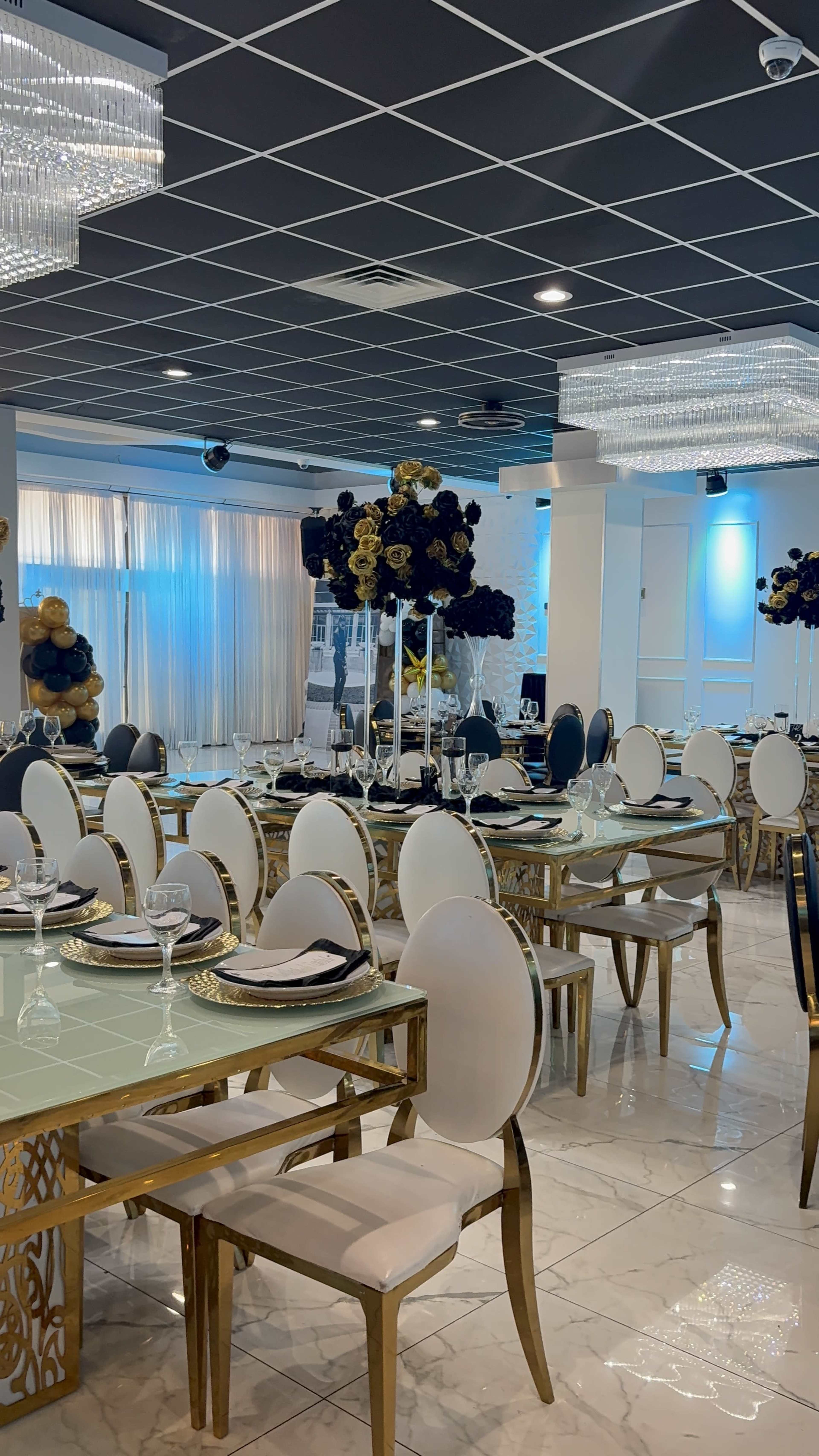 The image shows a modern dining area with elegantly set tables, white chairs, and decorative centerpieces featuring dark flowers against a backdrop of soft lighting and chandeliers.