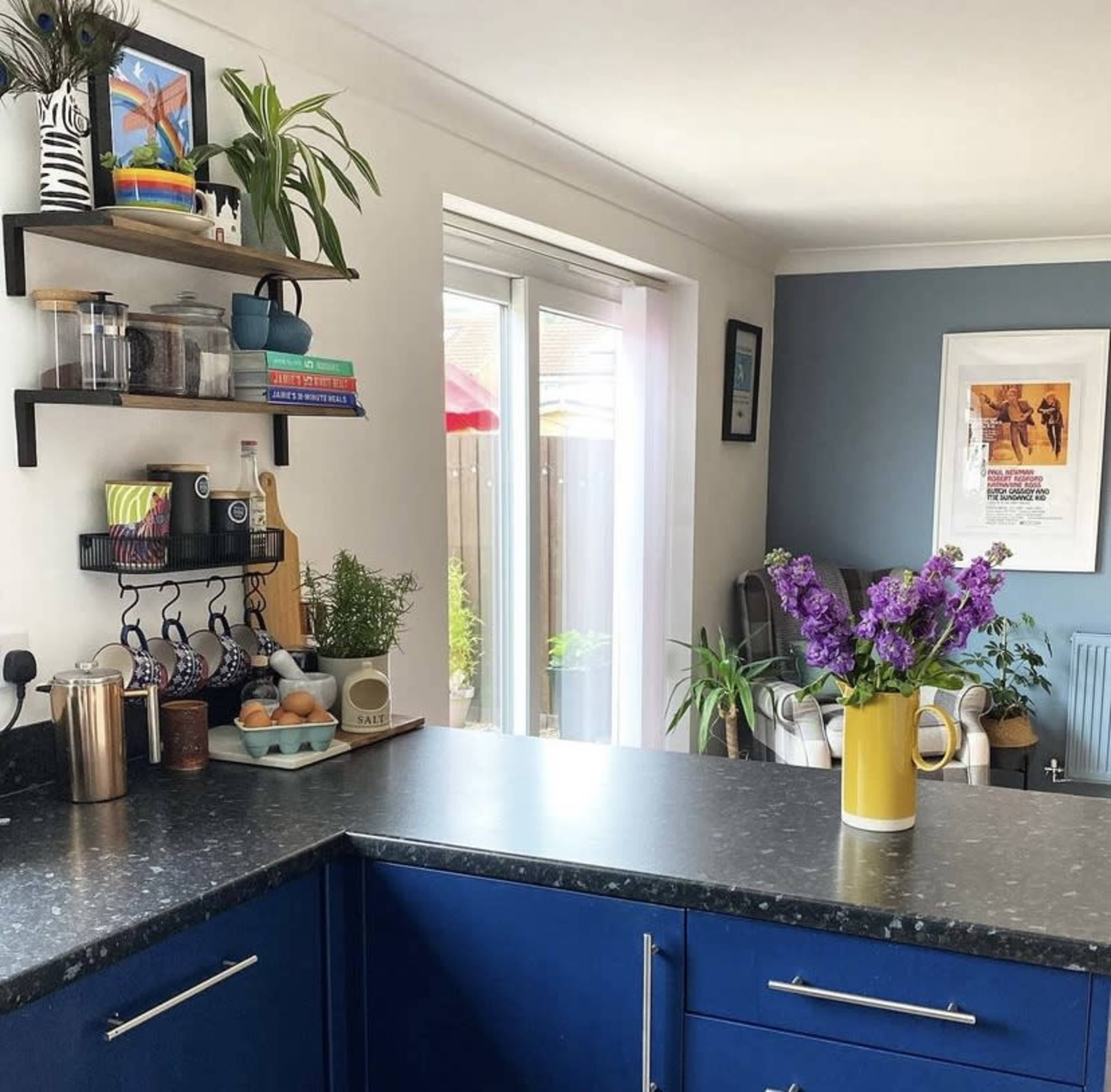 A modern kitchen features a blue countertop, shelves with various kitchen items, and a vase of purple flowers.