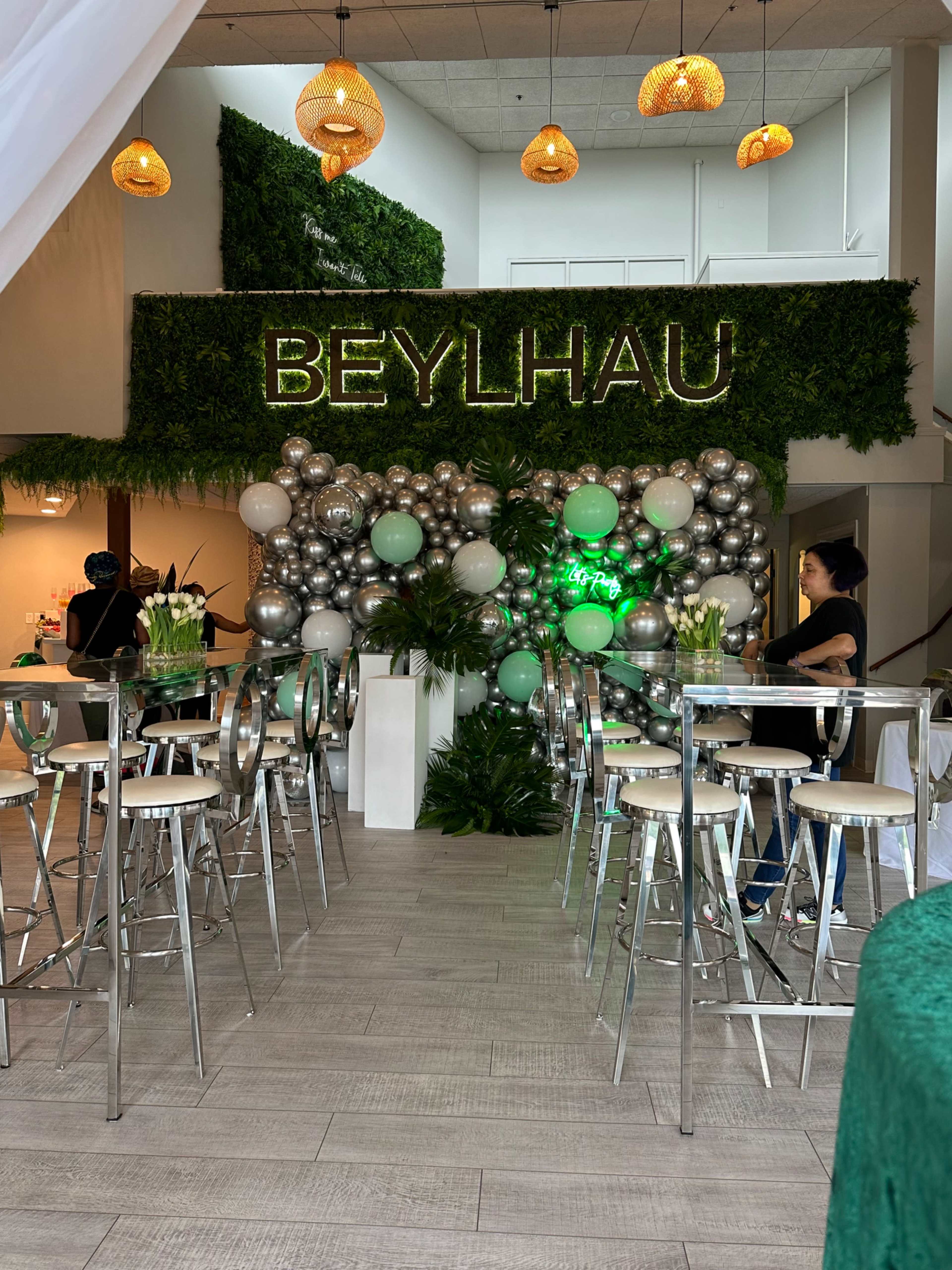 An event space decorated with a greenery wall and a balloon arrangement, featuring a sign that reads "BEYLHAU," along with transparent bar stools and tables.