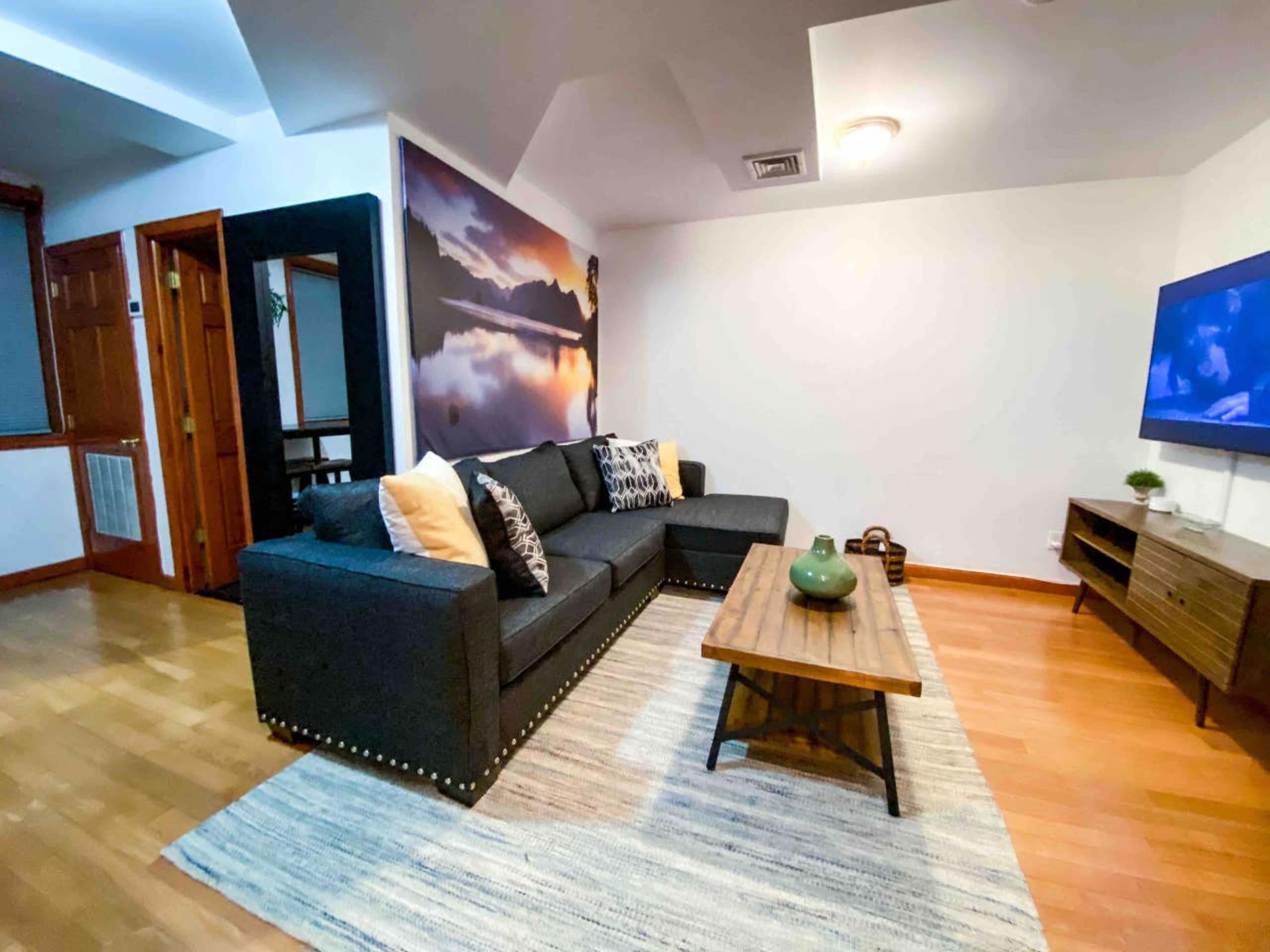A contemporary living room with a dark sectional sofa, a wooden coffee table, and a large wall-mounted TV, complemented by a wall art piece and wooden flooring.