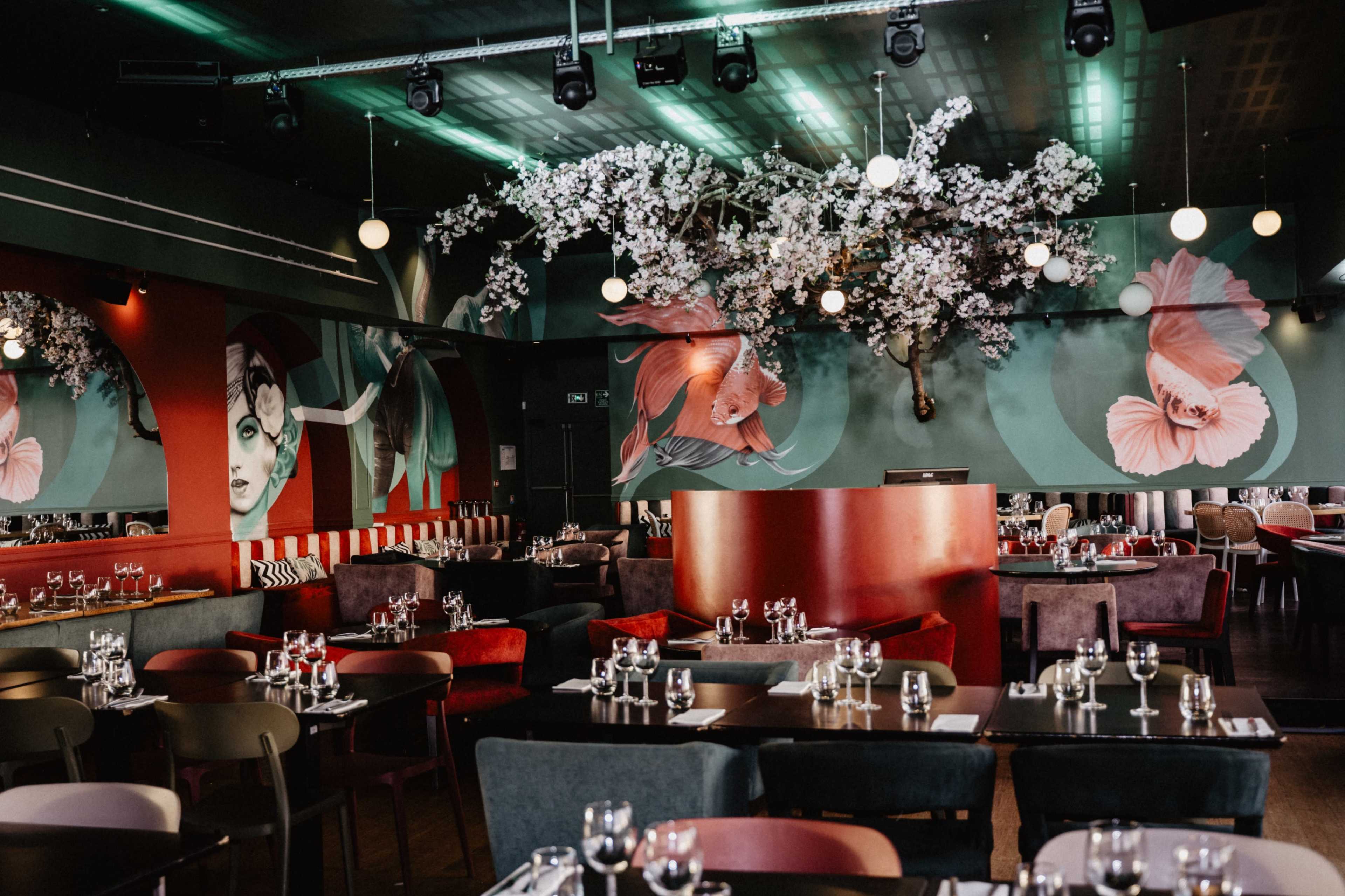 The image shows a modern restaurant interior featuring decorative floral murals, ornate lighting, and elegantly arranged dining tables with glassware.