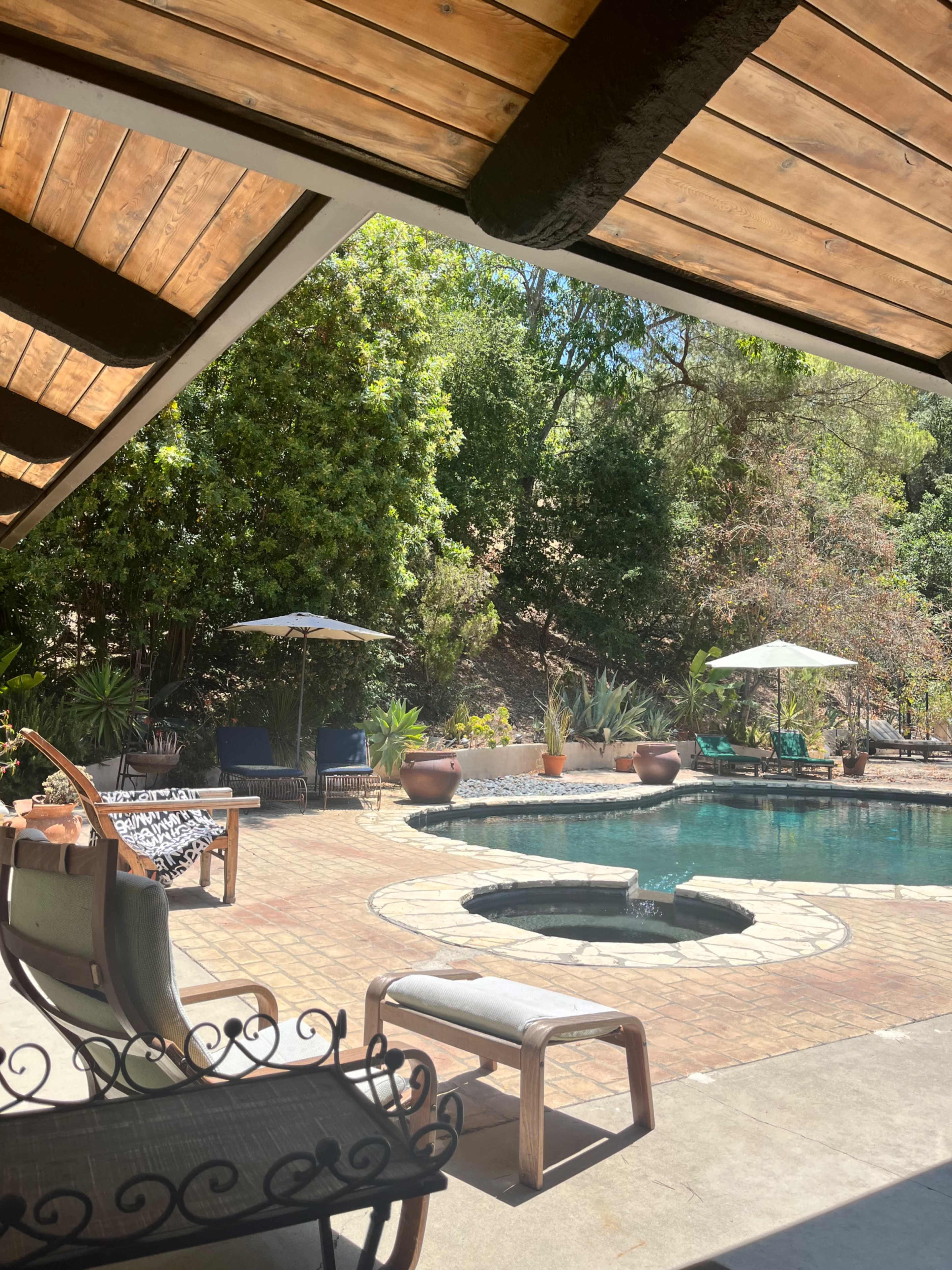 A patio with a swimming pool, surrounded by lounge chairs, umbrellas, and landscaping, is visible underneath a wooden roof structure.