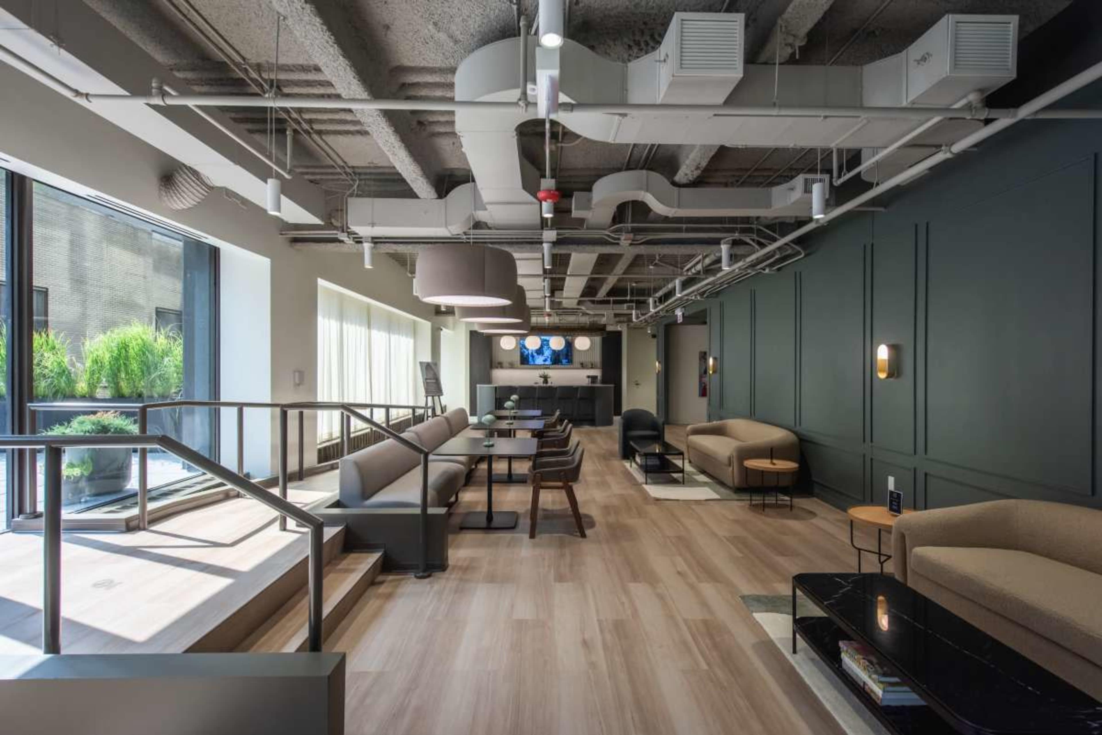 The image depicts a modern interior space featuring a seating area with sofas, tables, and a light color scheme, complemented by large windows and exposed ceiling ducts.