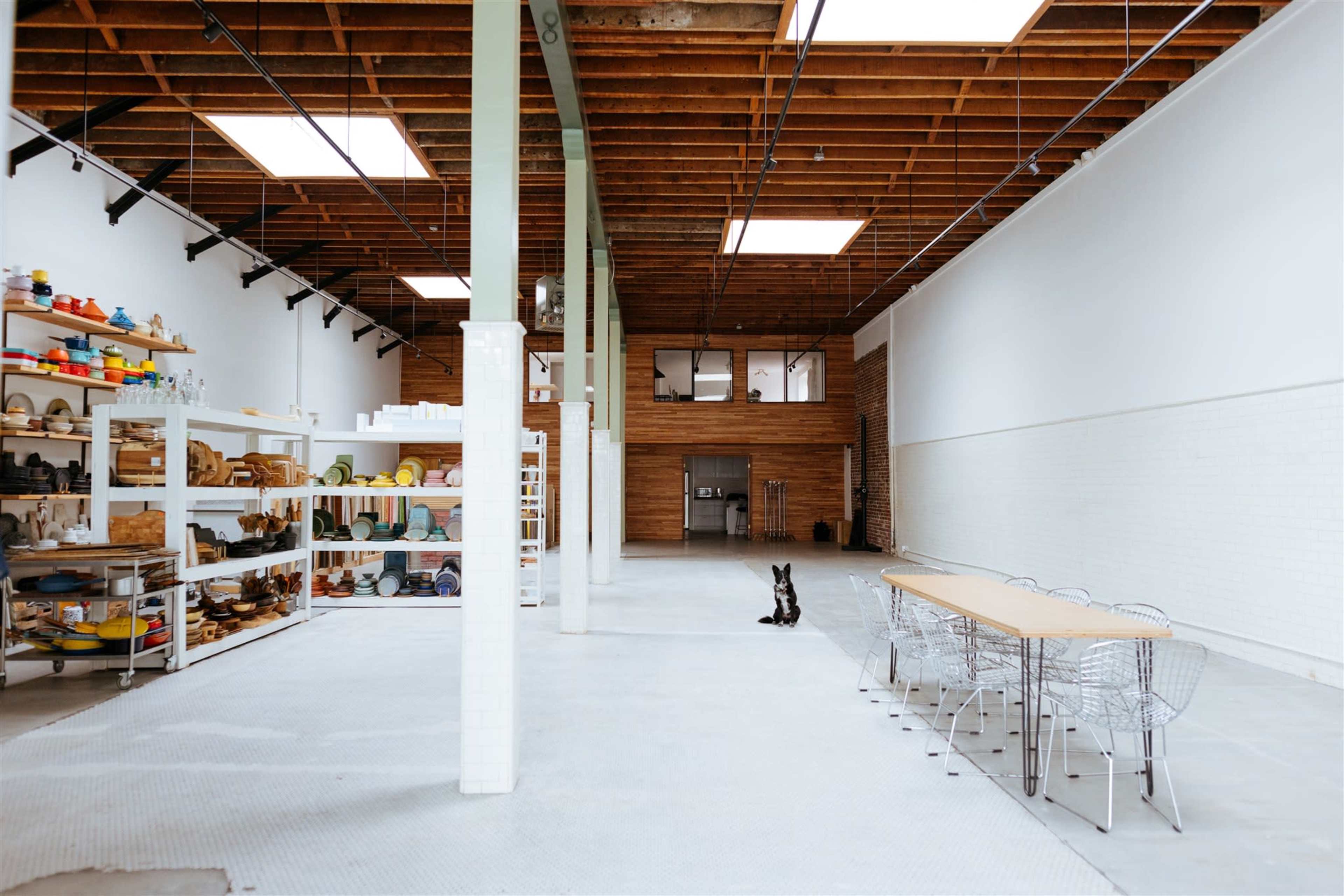 A spacious, well-lit interior features a long wooden table, a collection of colorful pottery on shelves, and a dog sitting on the concrete floor.