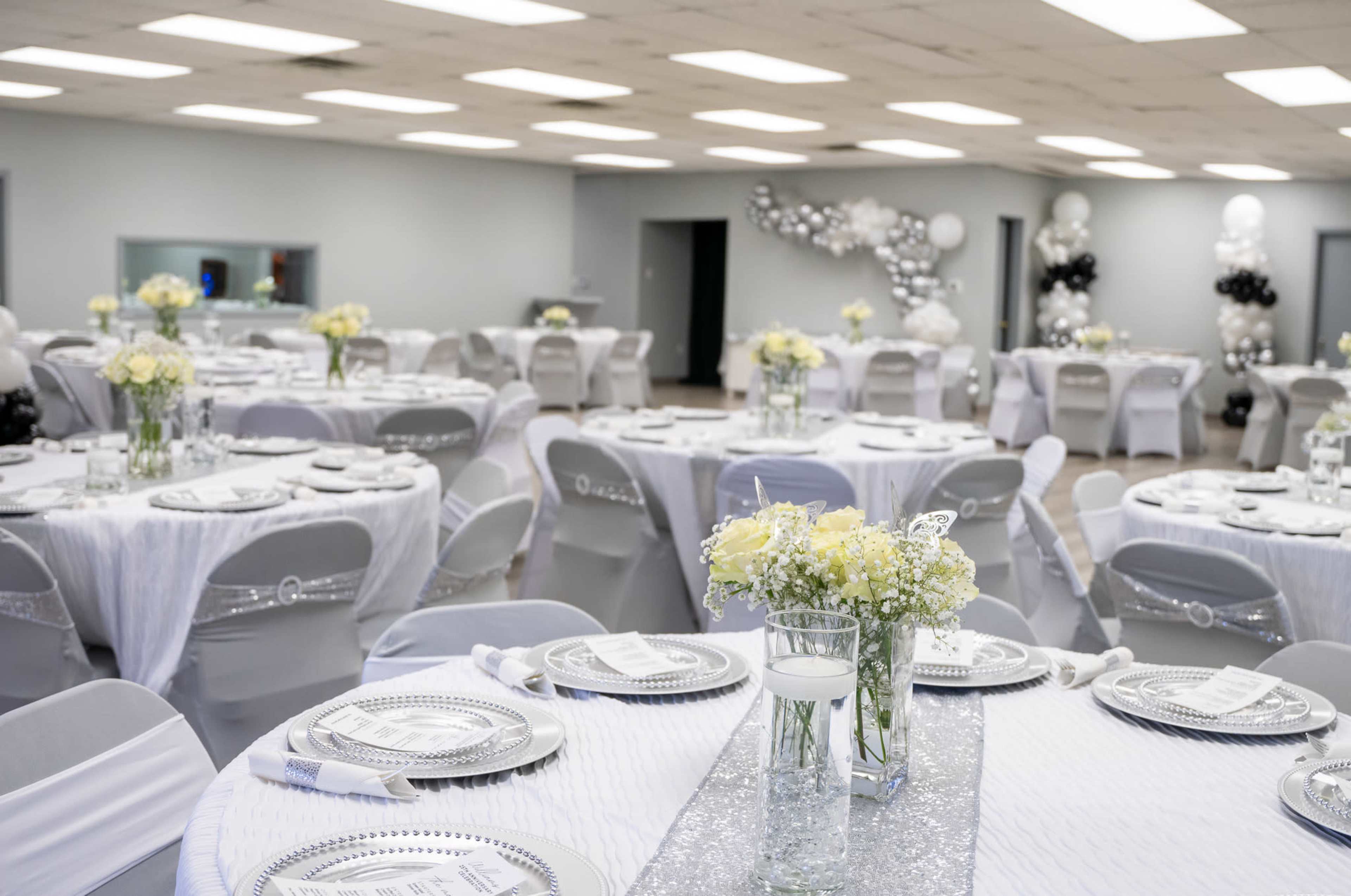 The image shows a banquet hall set up for an event with round tables covered in white and silver tablecloths, adorned with floral centerpieces and decorative place settings.