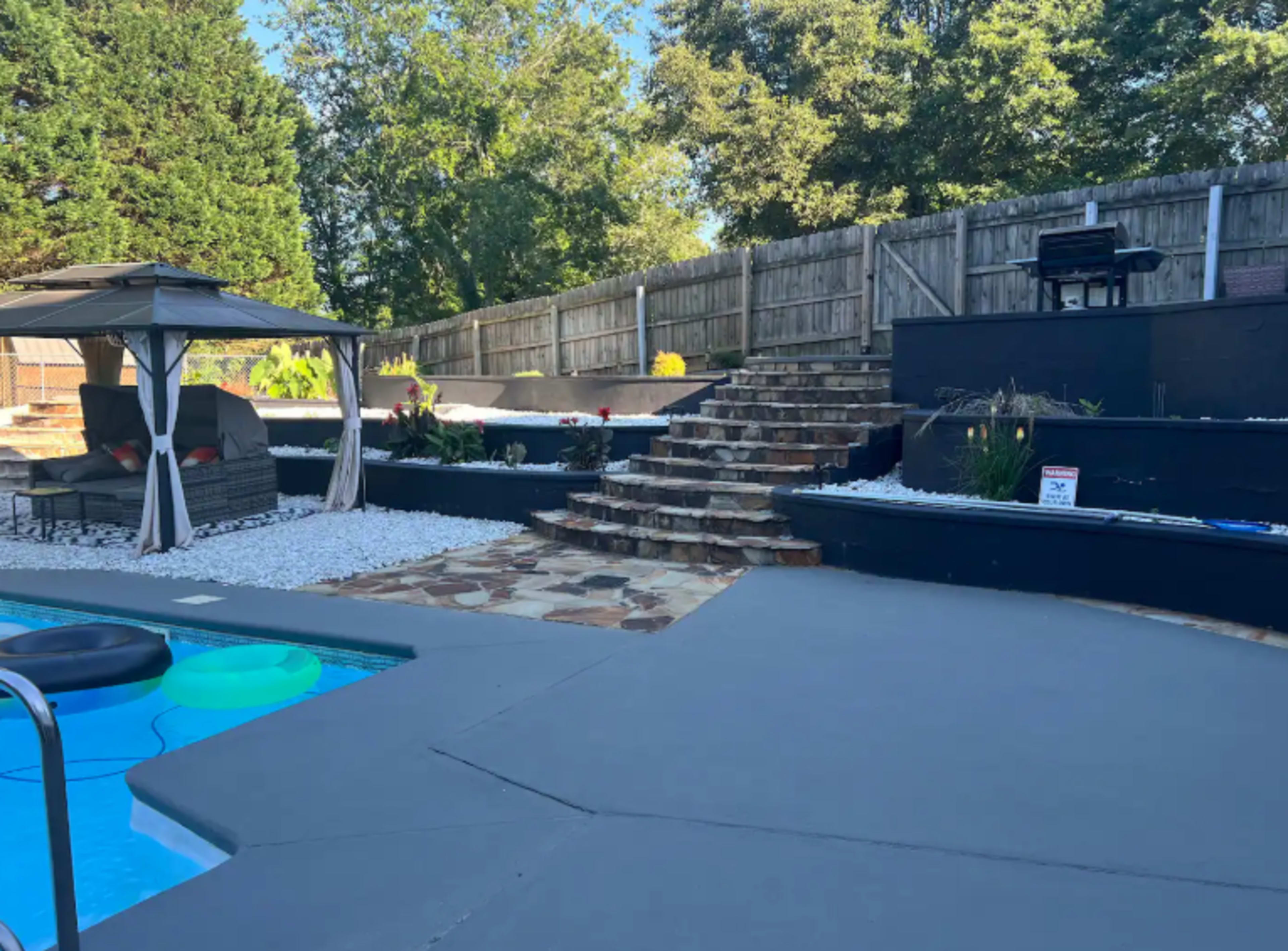 The image shows a backyard with a swimming pool, a gazebo, stone steps leading to an elevated area, and landscaped sections featuring grass and decorative stones.