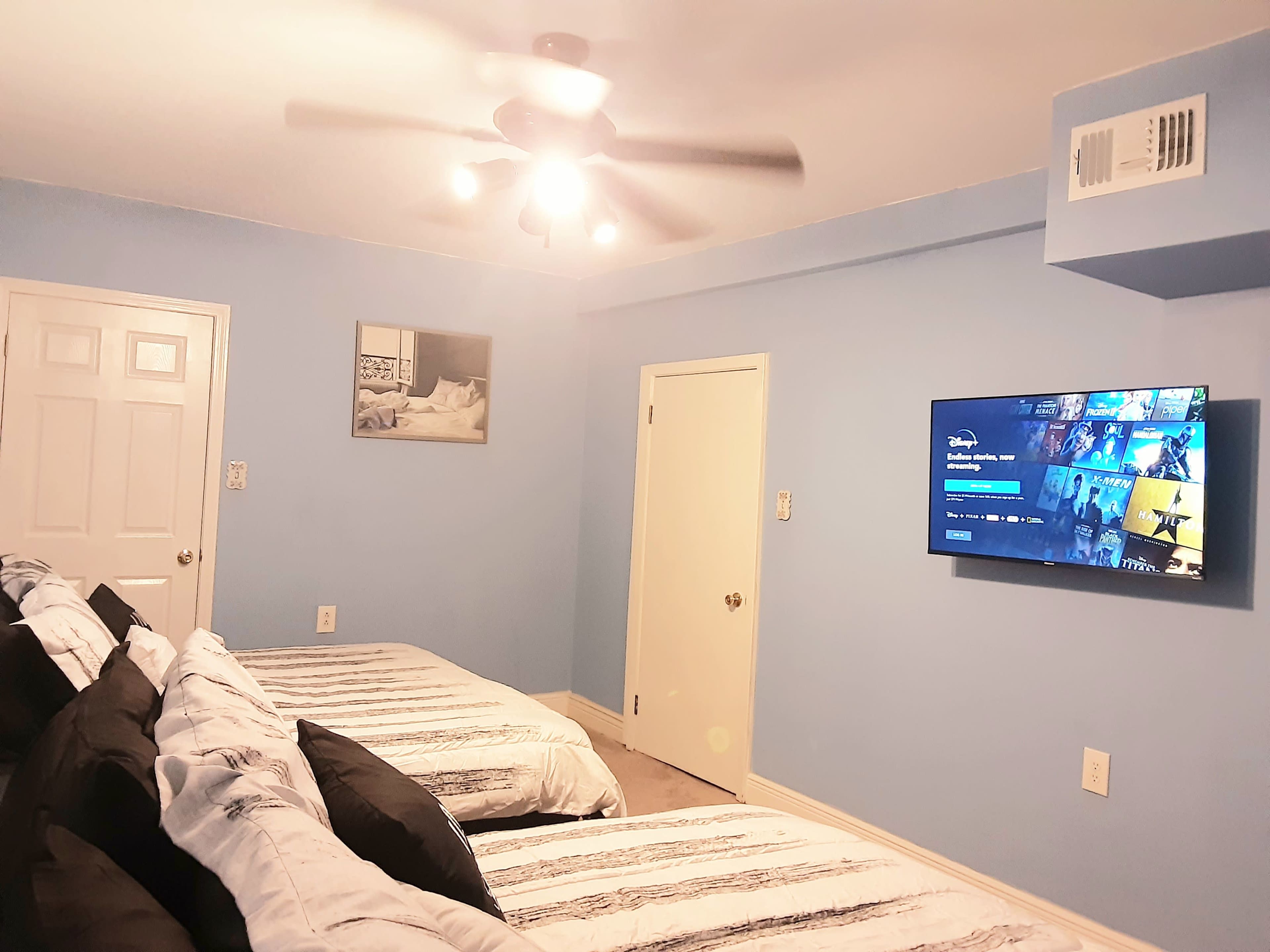 A bedroom with two beds, a mounted television displaying a streaming service, and light blue walls.
