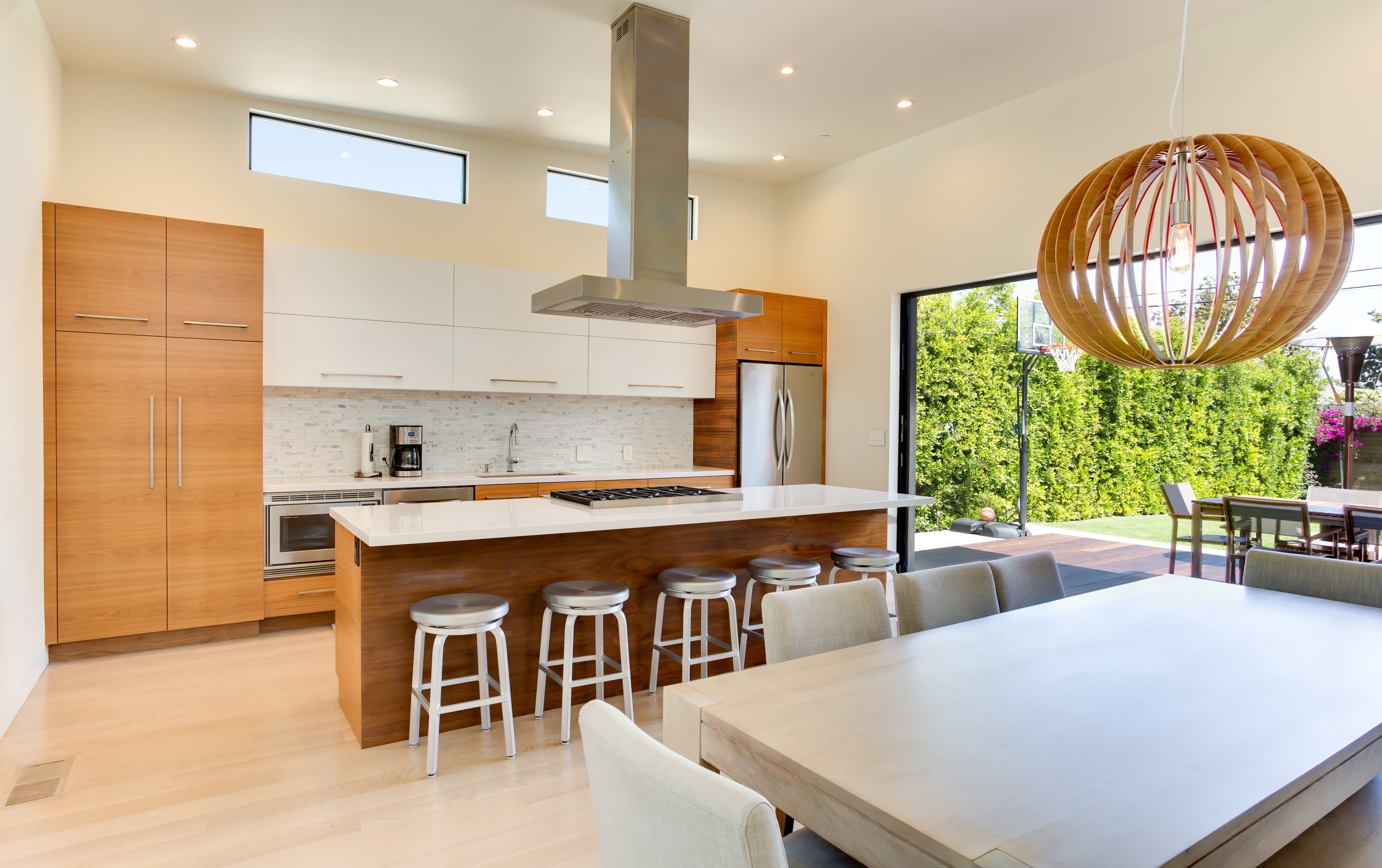 The image shows a modern kitchen with a central island, sleek cabinetry, and a dining area that opens to an outdoor view.