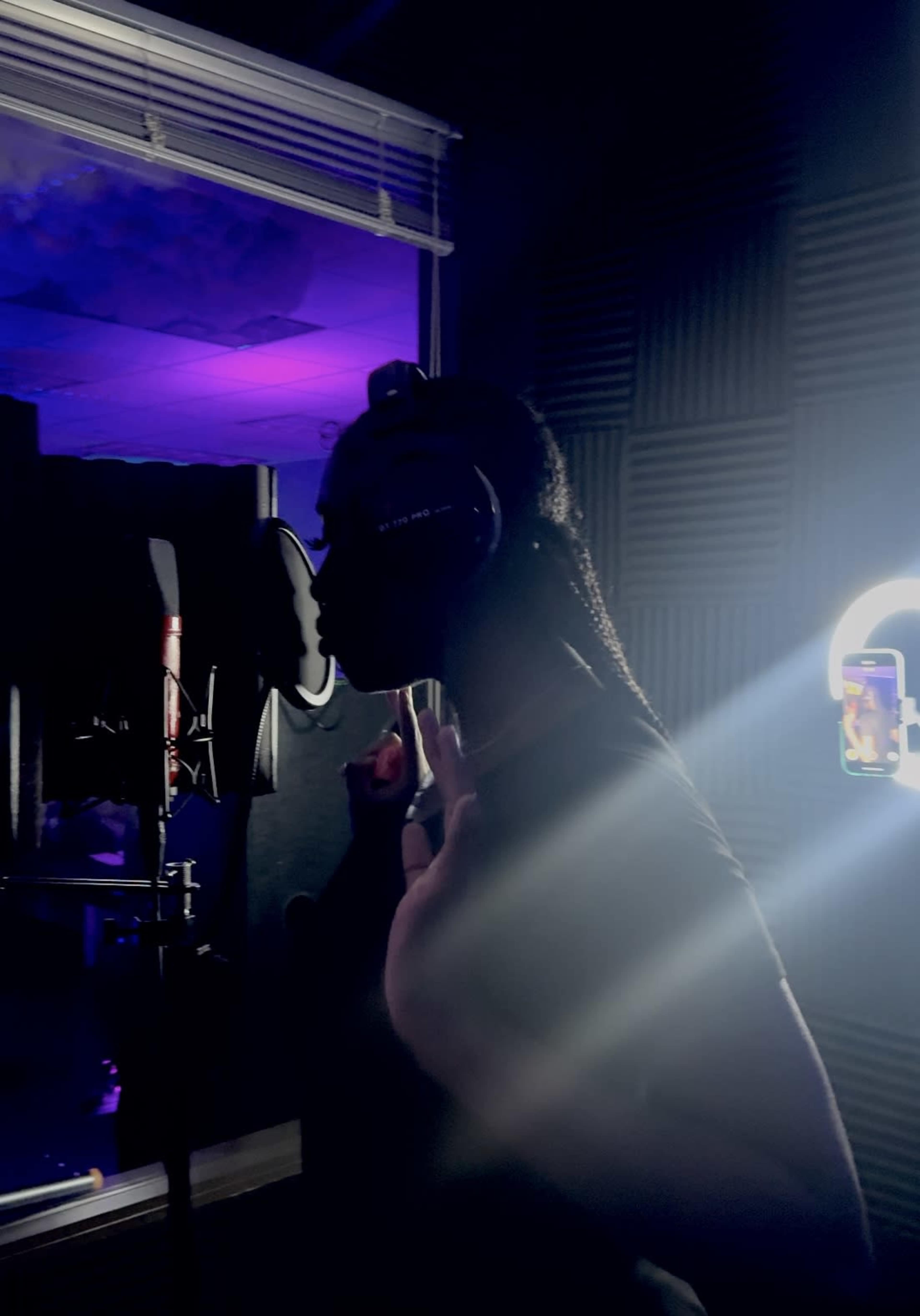 A person with braided hair stands in front of a microphone in a dimly lit recording studio, wearing headphones and gesturing with one hand.