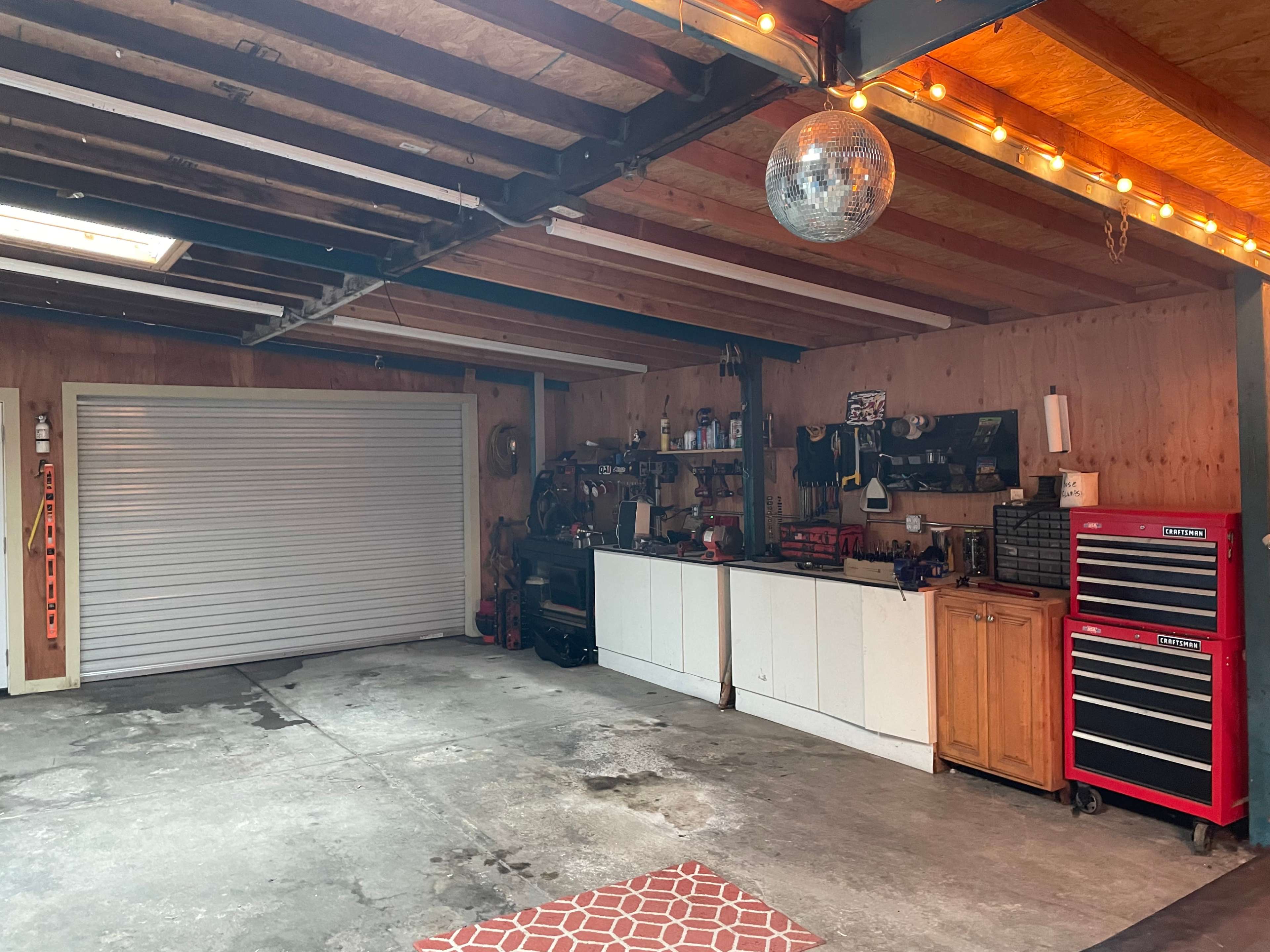 A garage with a roll-up door, a workbench against one wall, and two tool chests, all illuminated by string lights overhead.
