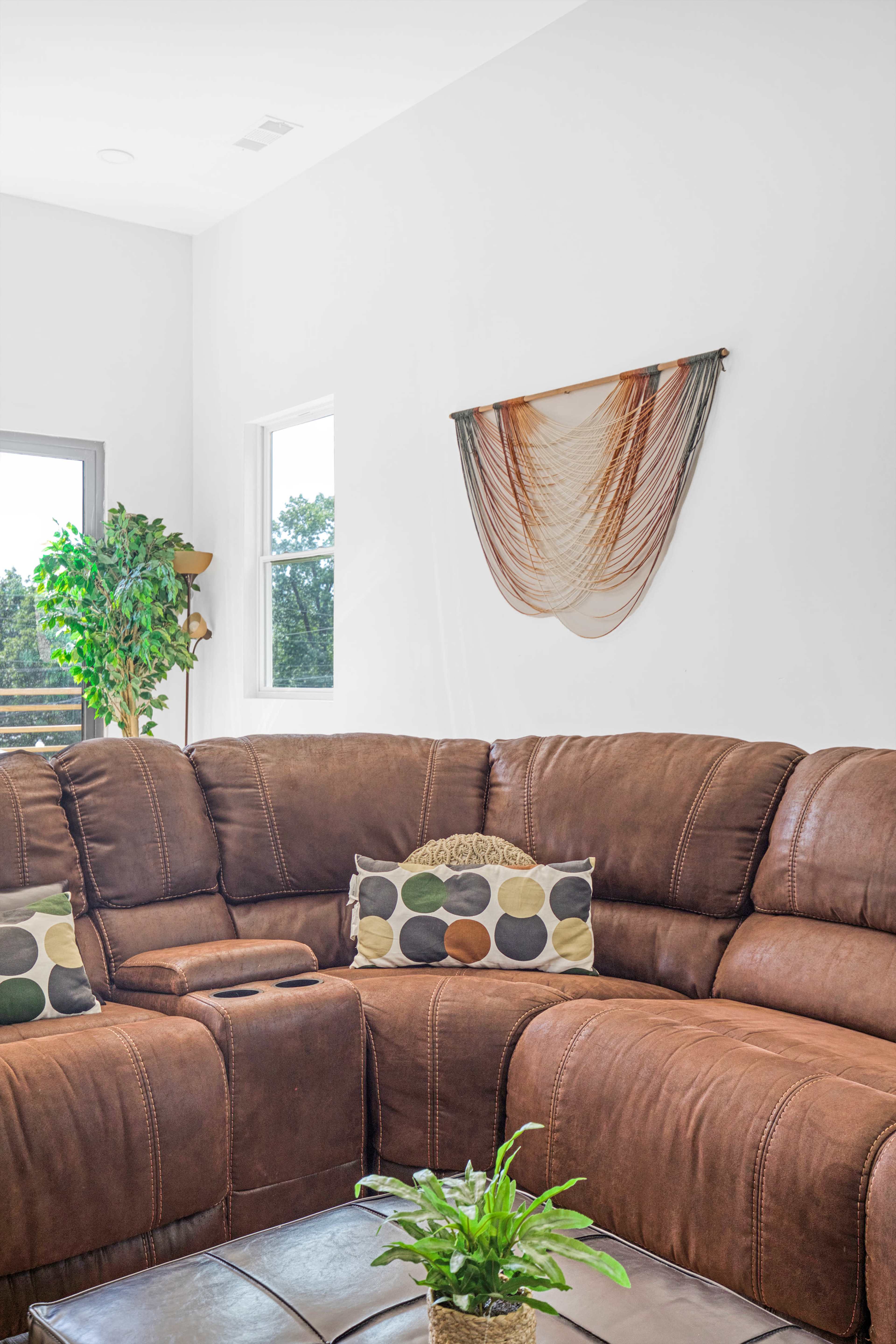 A brown leather sectional sofa is positioned in a bright living room with a green plant and decorative pillows on it, alongside a wall featuring a cloth hanging.