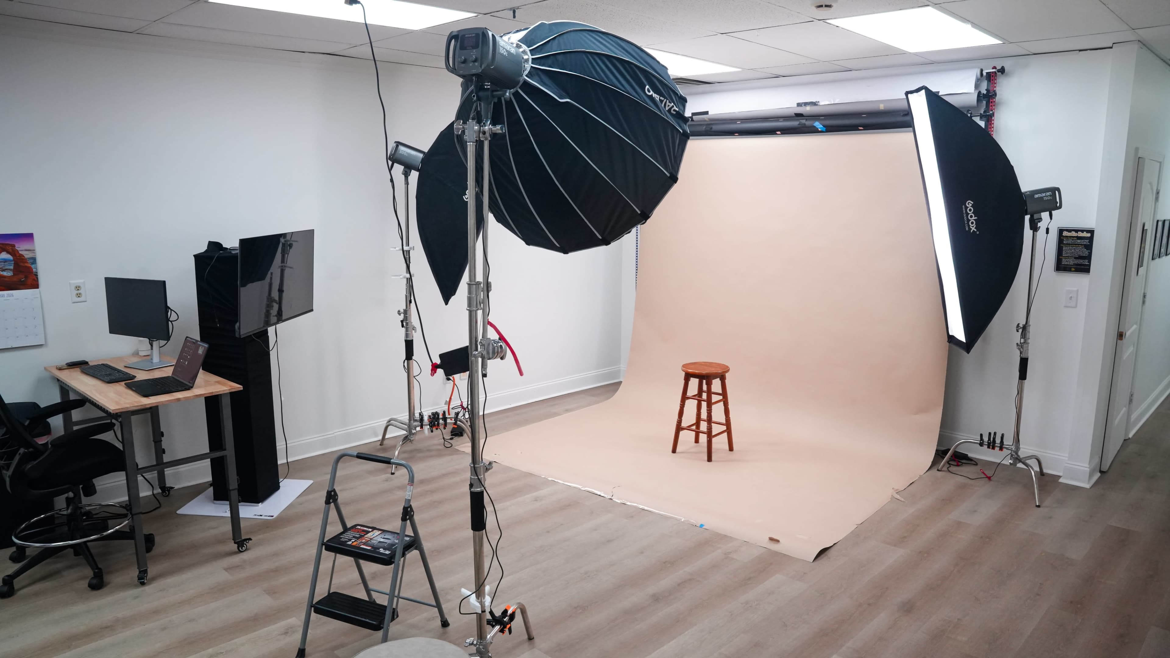 The image shows a photography studio setup with a stool in front of a beige backdrop, flanked by large softbox lights and equipment on stands.
