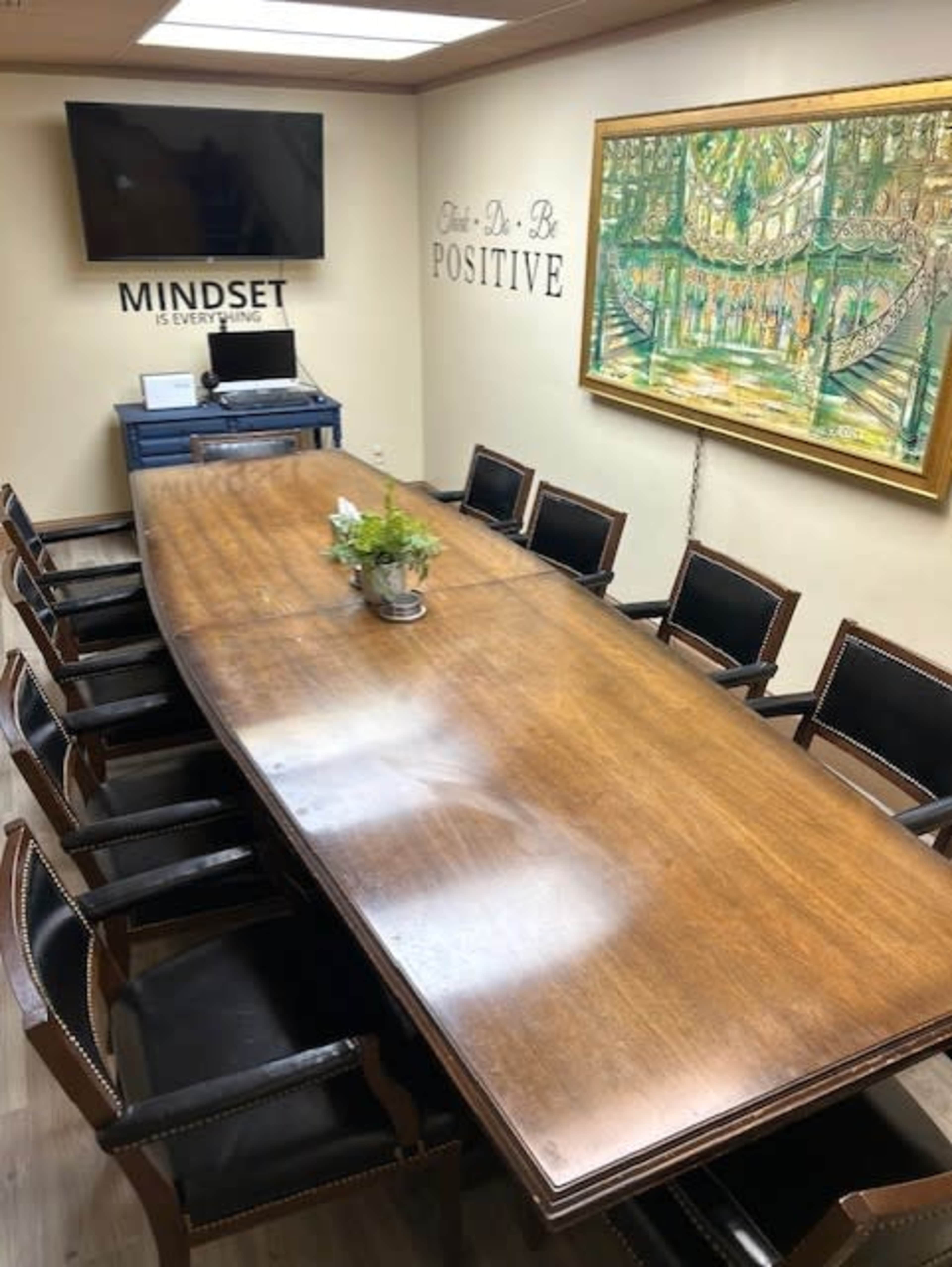 A long oval conference table surrounded by black chairs is situated in a meeting room with motivational wall art and a large painting.