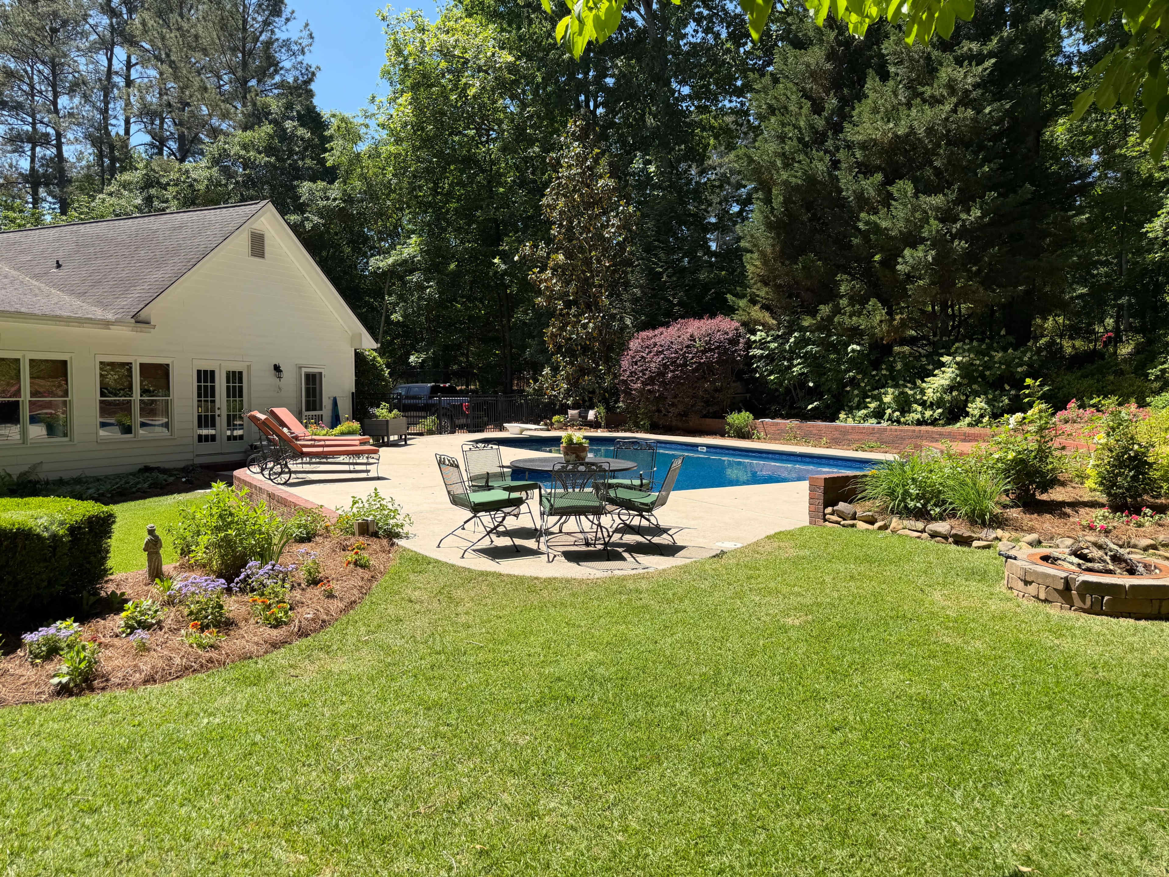 A well-maintained backyard features a pool surrounded by lounge chairs and a patio set, with lush greenery and flowers enhancing the landscape.