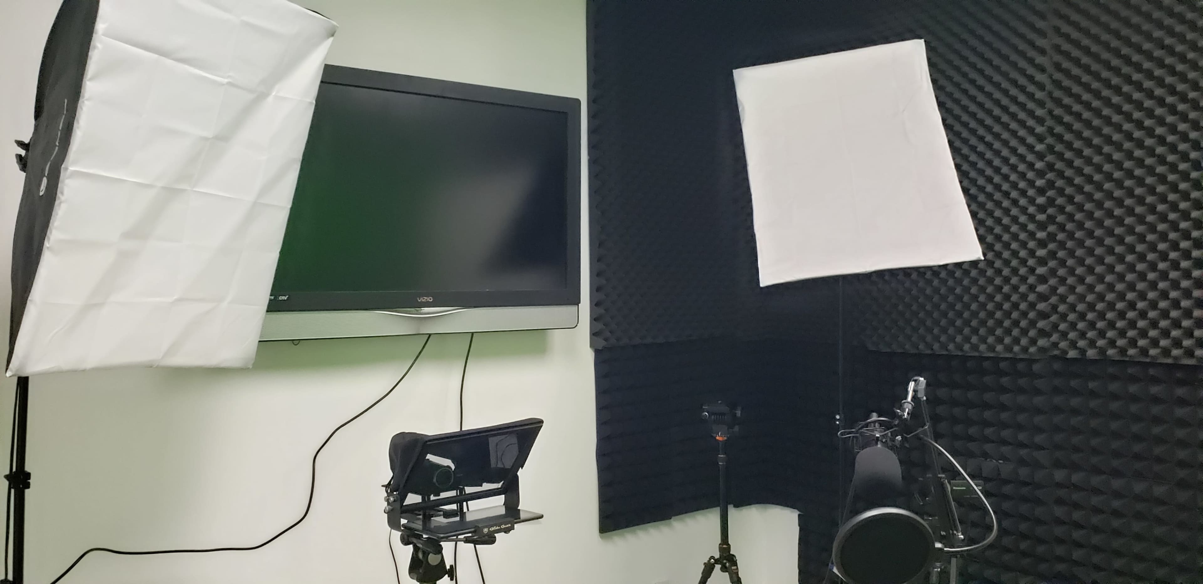 A studio setup featuring a flat-screen TV, two softbox lights, a camera on a tripod, and soundproofing panels on the walls.