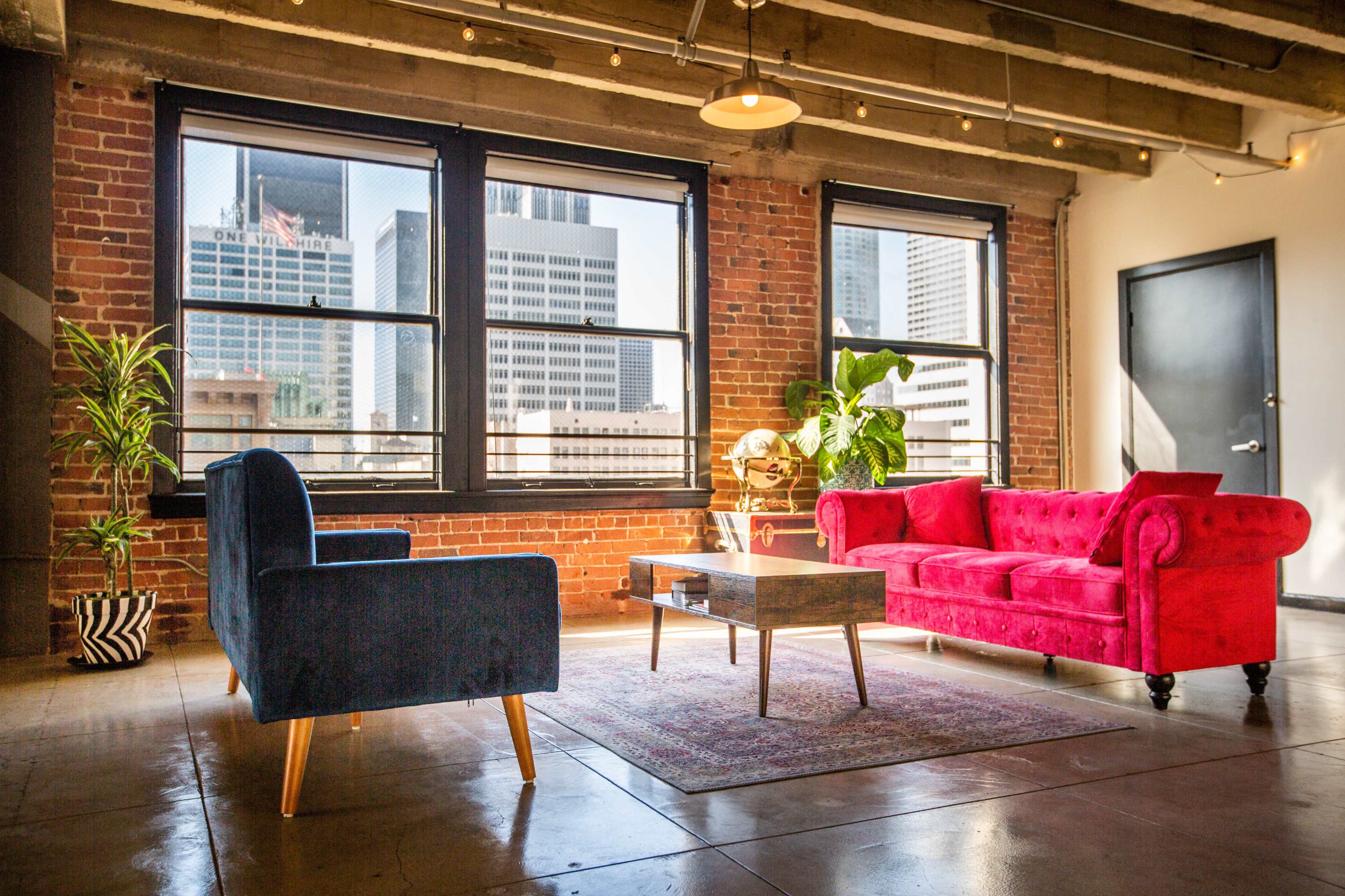 The image shows a modern living area featuring a red sofa, a blue chair, a wooden coffee table, and large windows with city views.