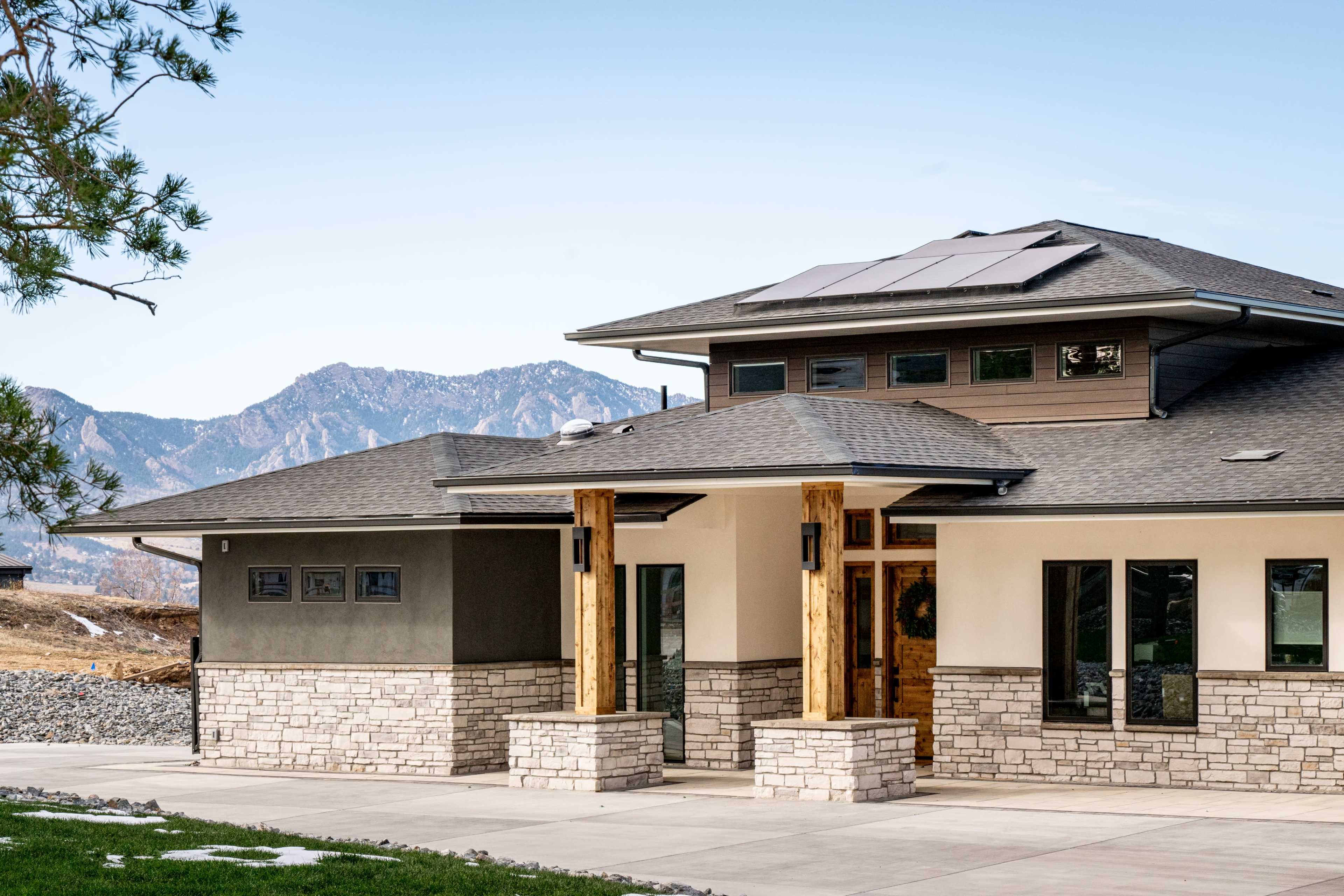 A modern house with a combination of stone and stucco exterior features a solar panel roof and is set against a backdrop of mountains.