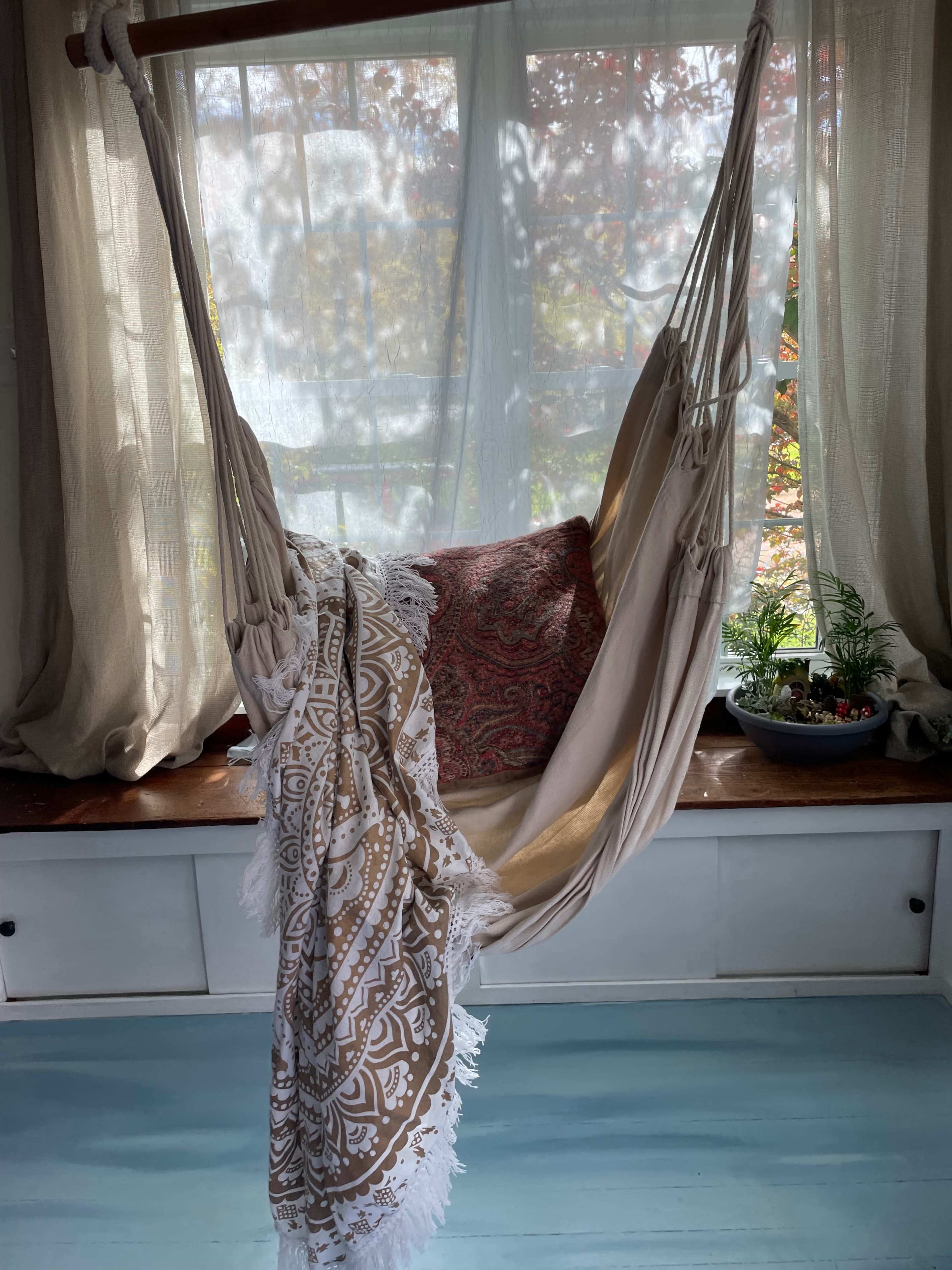 A lightweight hammock is suspended near a window, adorned with a patterned throw and cushion, in a sunlit room.