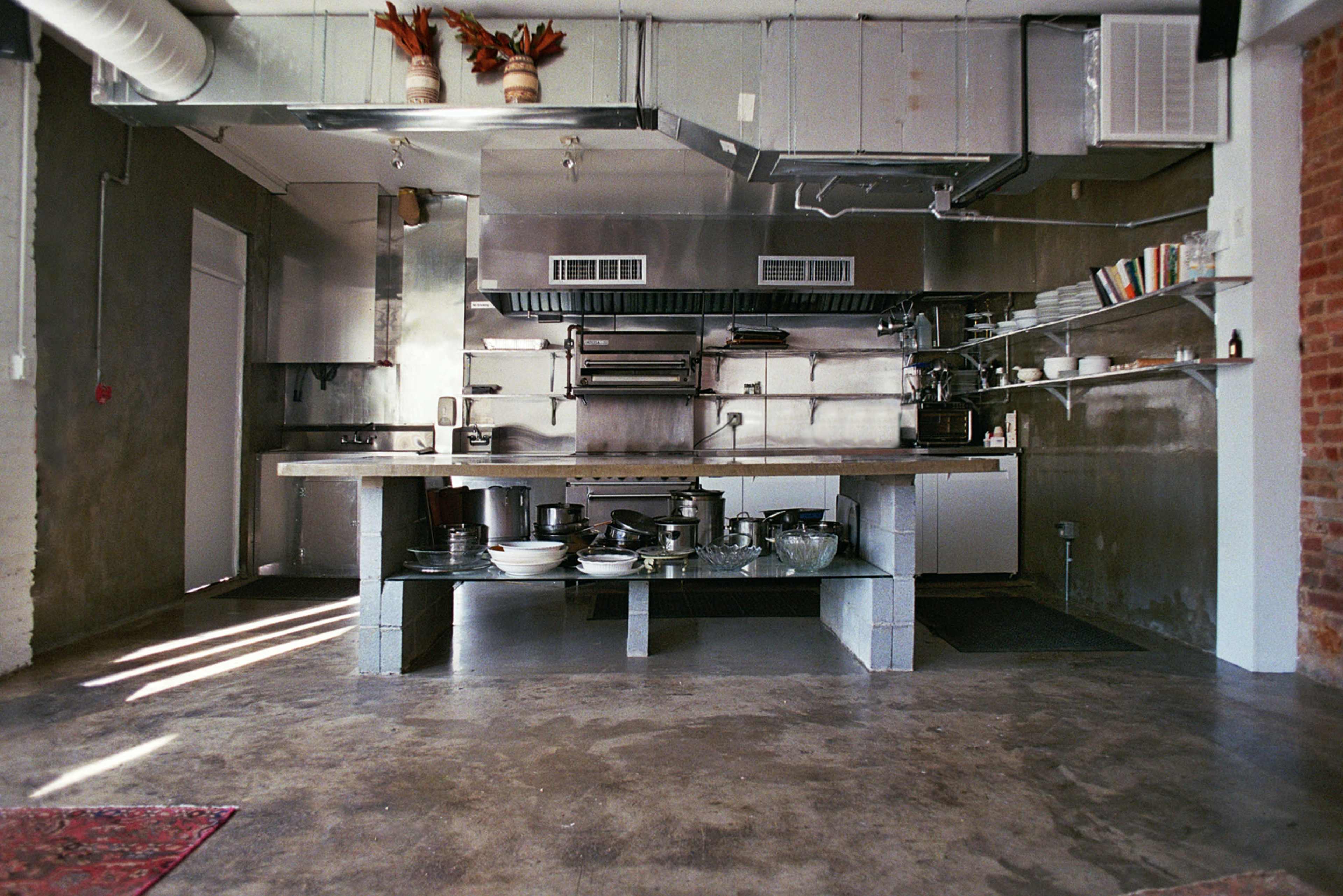 The image shows a modern kitchen with stainless steel countertops, an industrial stove, and various pots and utensils arranged on a concrete island.