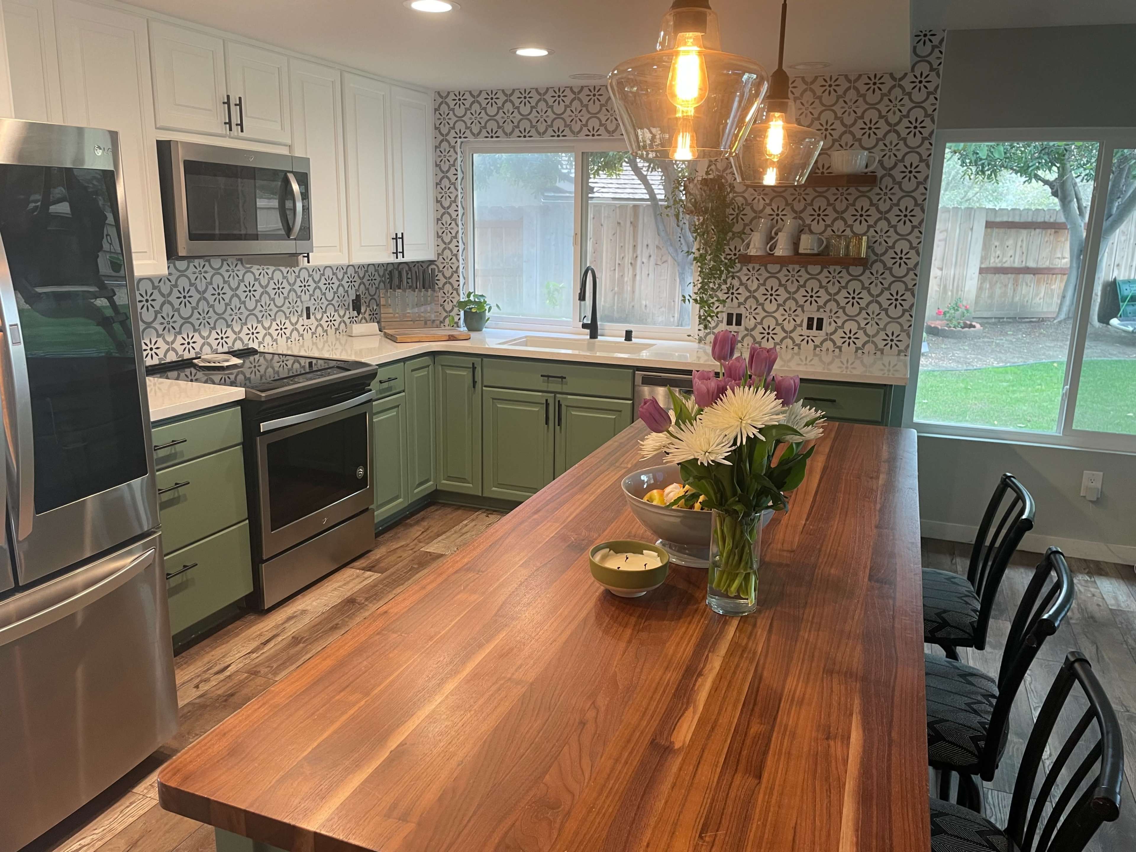 A modern kitchen with green cabinets, a large wooden island, a stainless steel fridge, and a window overlooking a backyard.