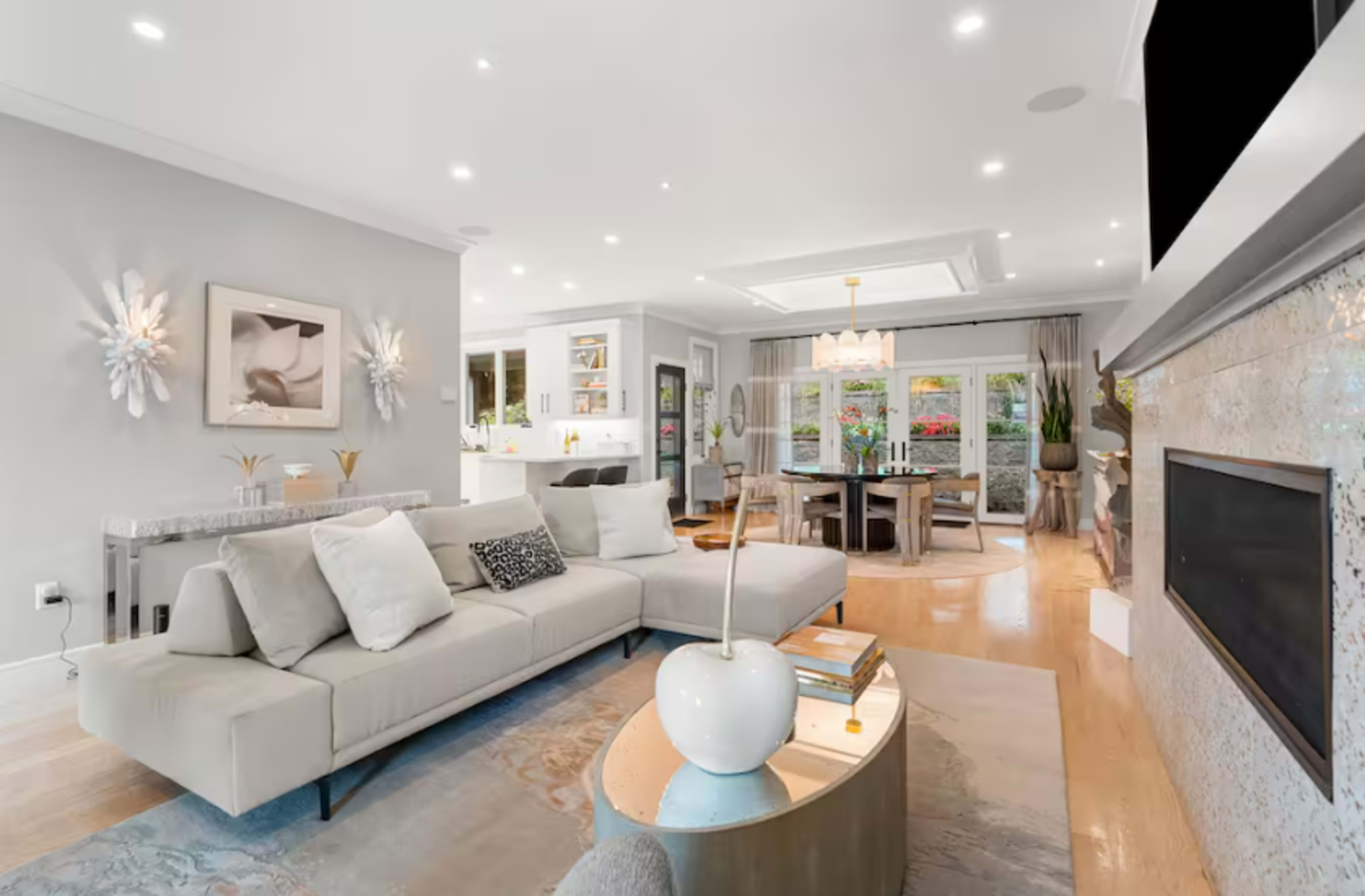 The living room features a light-colored sectional sofa, a round coffee table, and a dining area with a view of a garden through large windows.