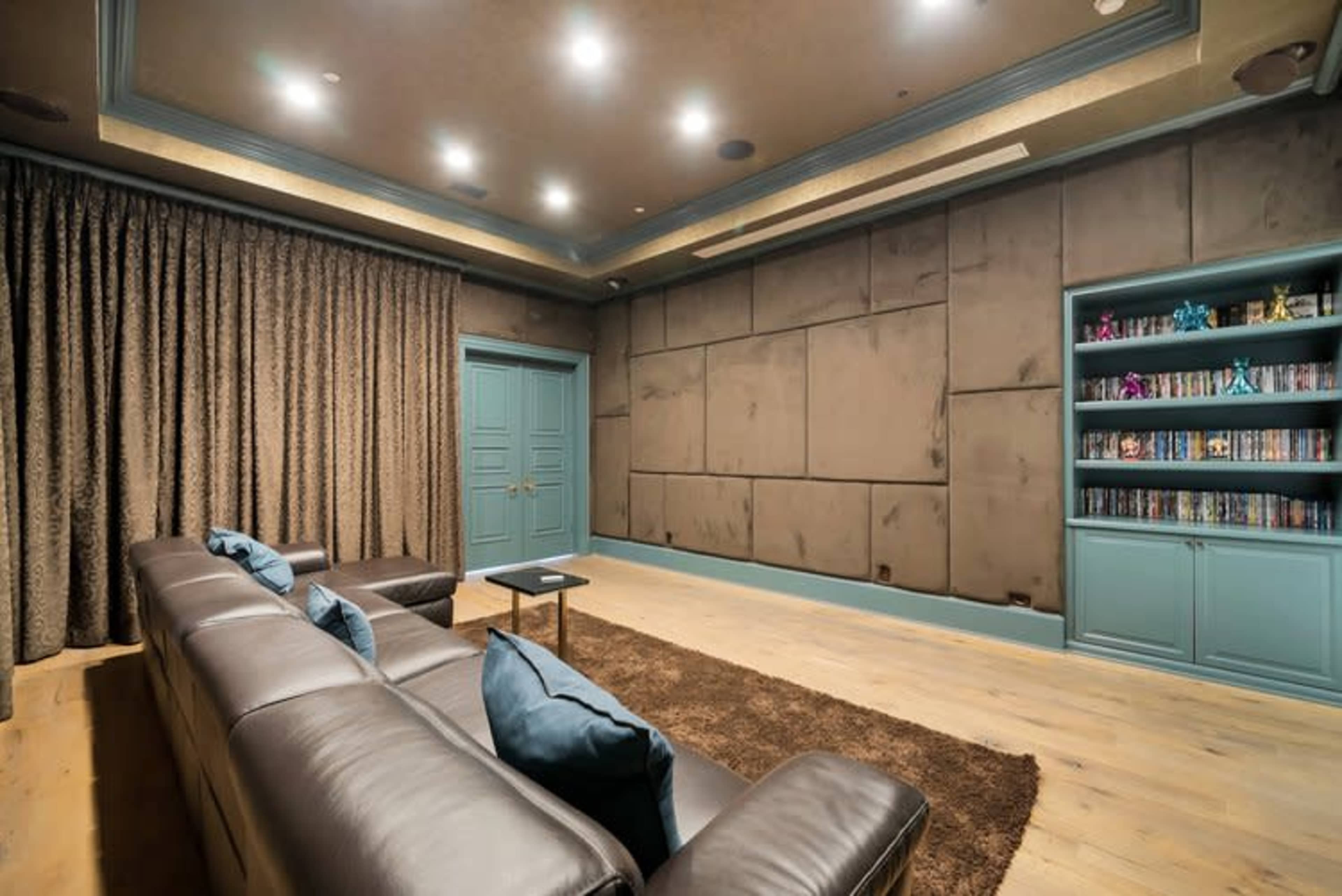 The image shows a cozy home theater with dark walls, a sectional leather sofa, and a bookshelf filled with DVDs.