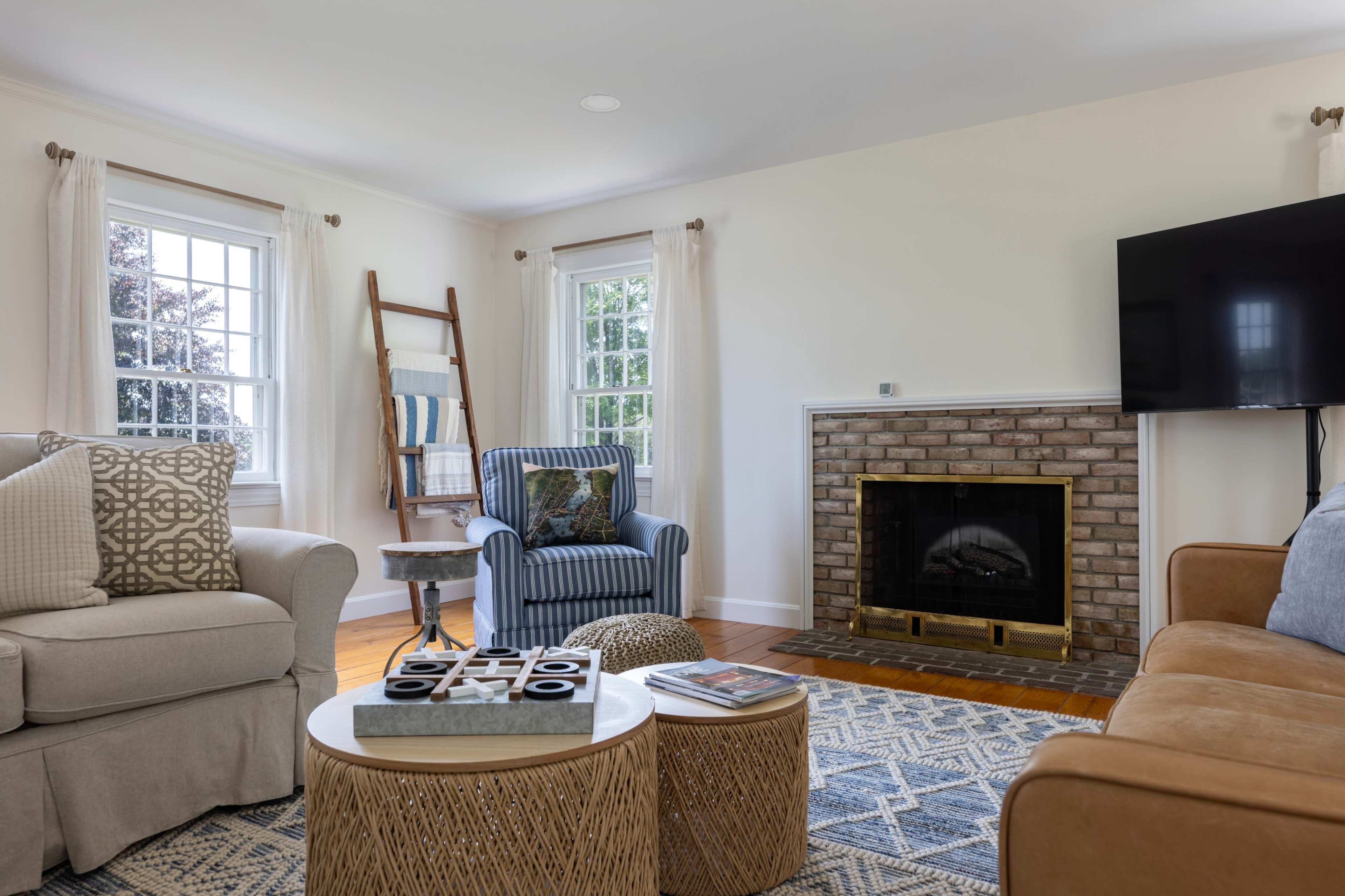 The image features a living room with a neutral-colored sofa, a brown leather chair, a round coffee table, and a brick fireplace, all arranged around a flat-screen TV mounted on the wall.