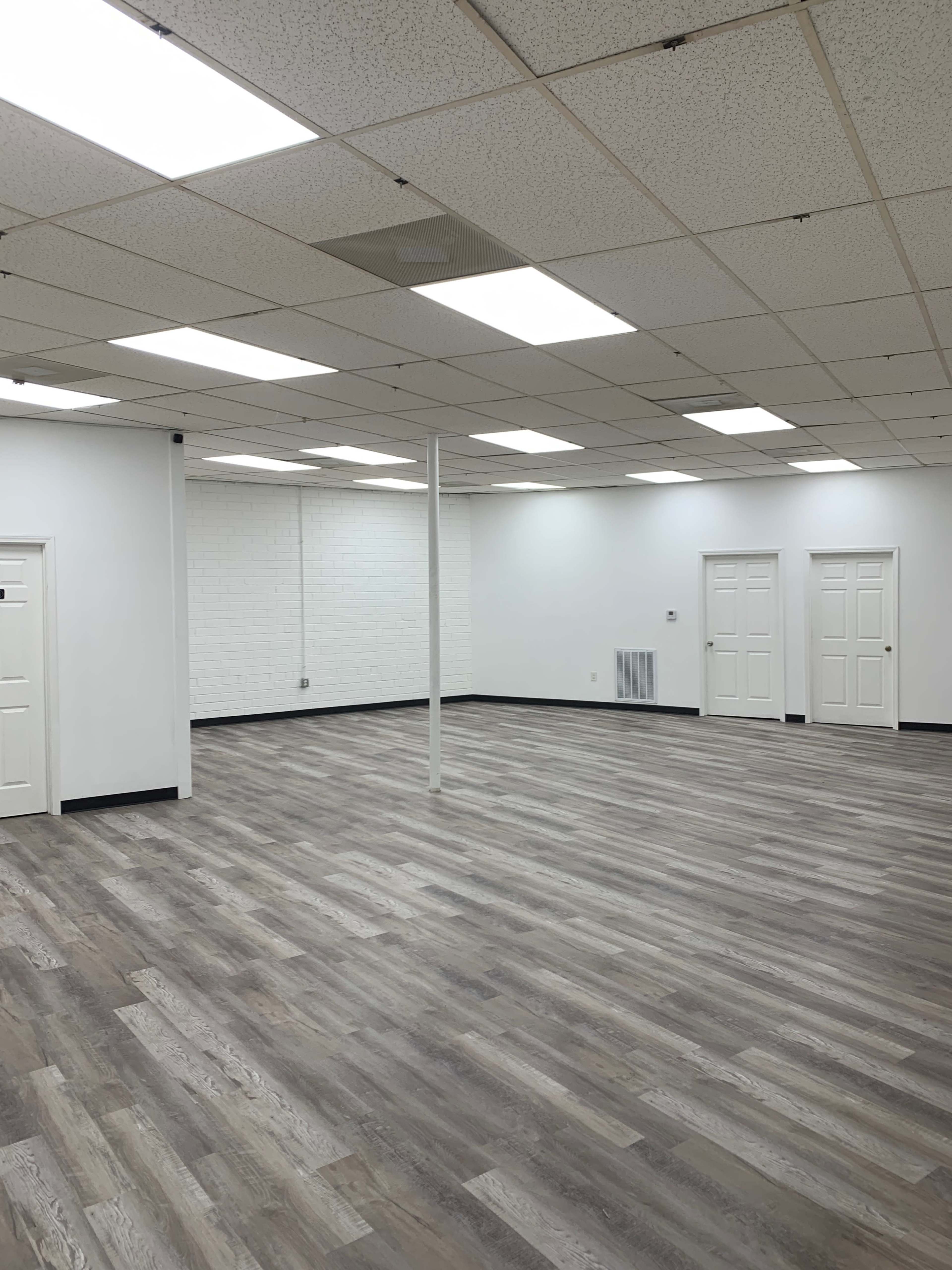 An empty, well-lit room with pale wooden flooring and white walls, featuring multiple doors and a central support column.