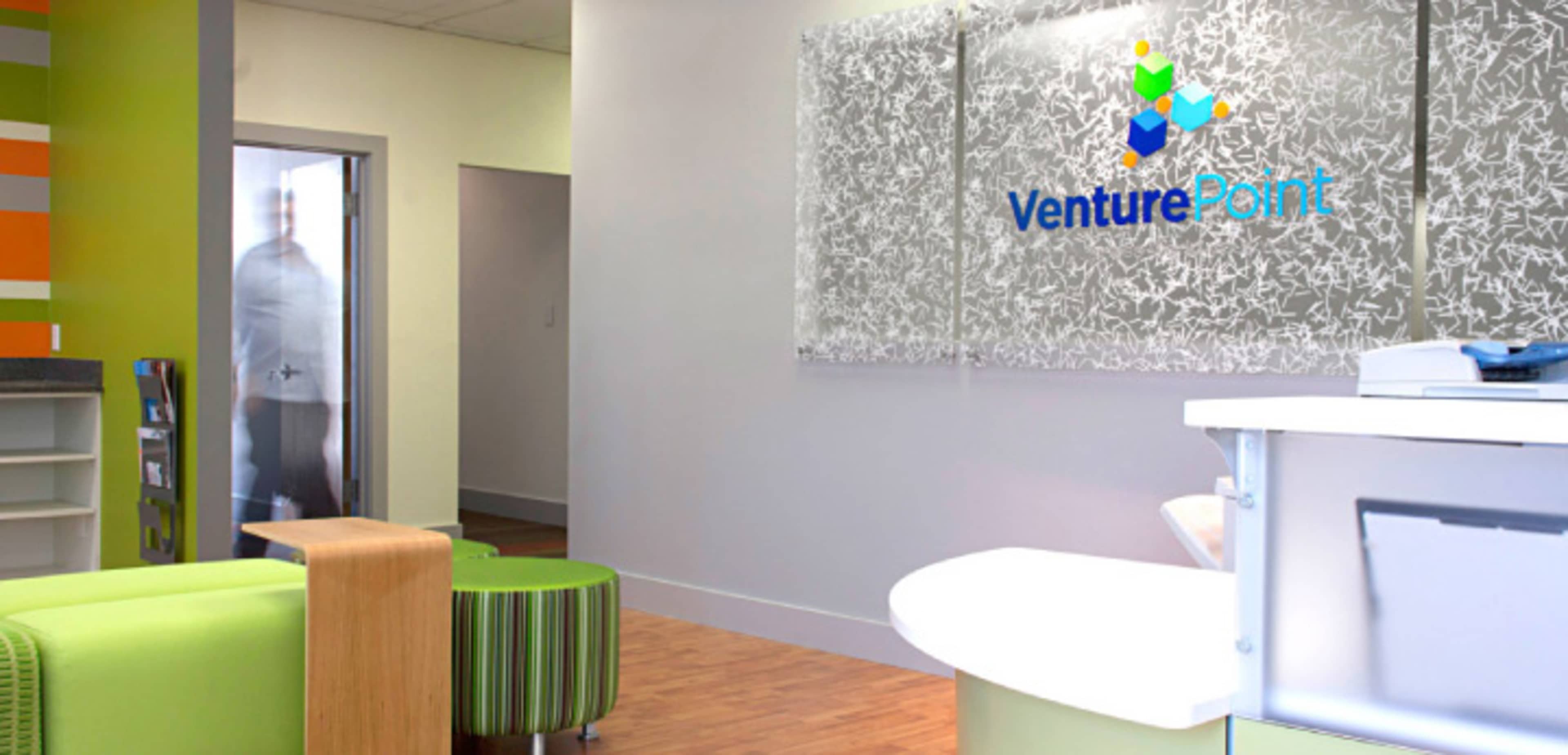 A modern reception area of an office with a logo display for VenturePoint on the wall, a circular white reception desk, and vibrant green seating.